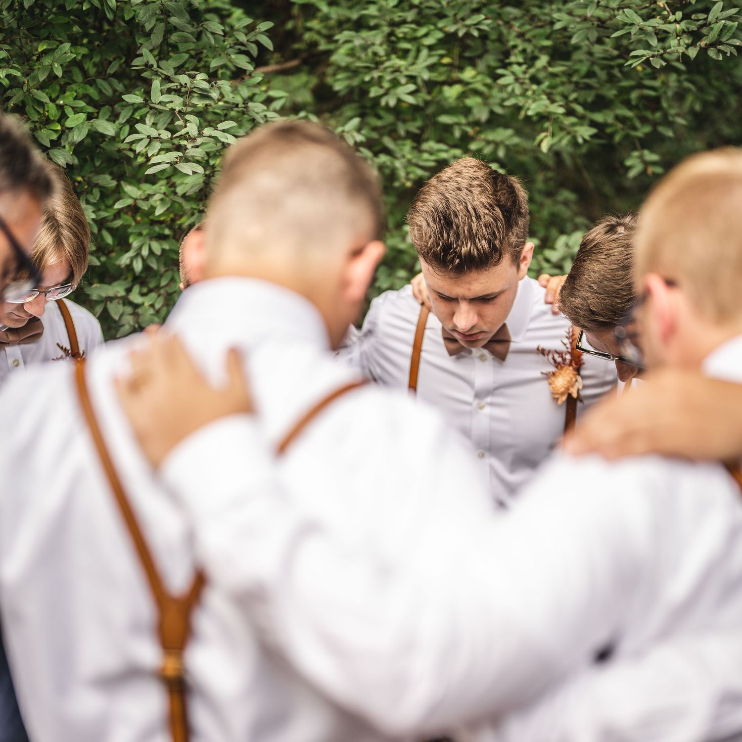 Wedding party in white shirts and leather suspenders gathered in intimate circle outdoors.