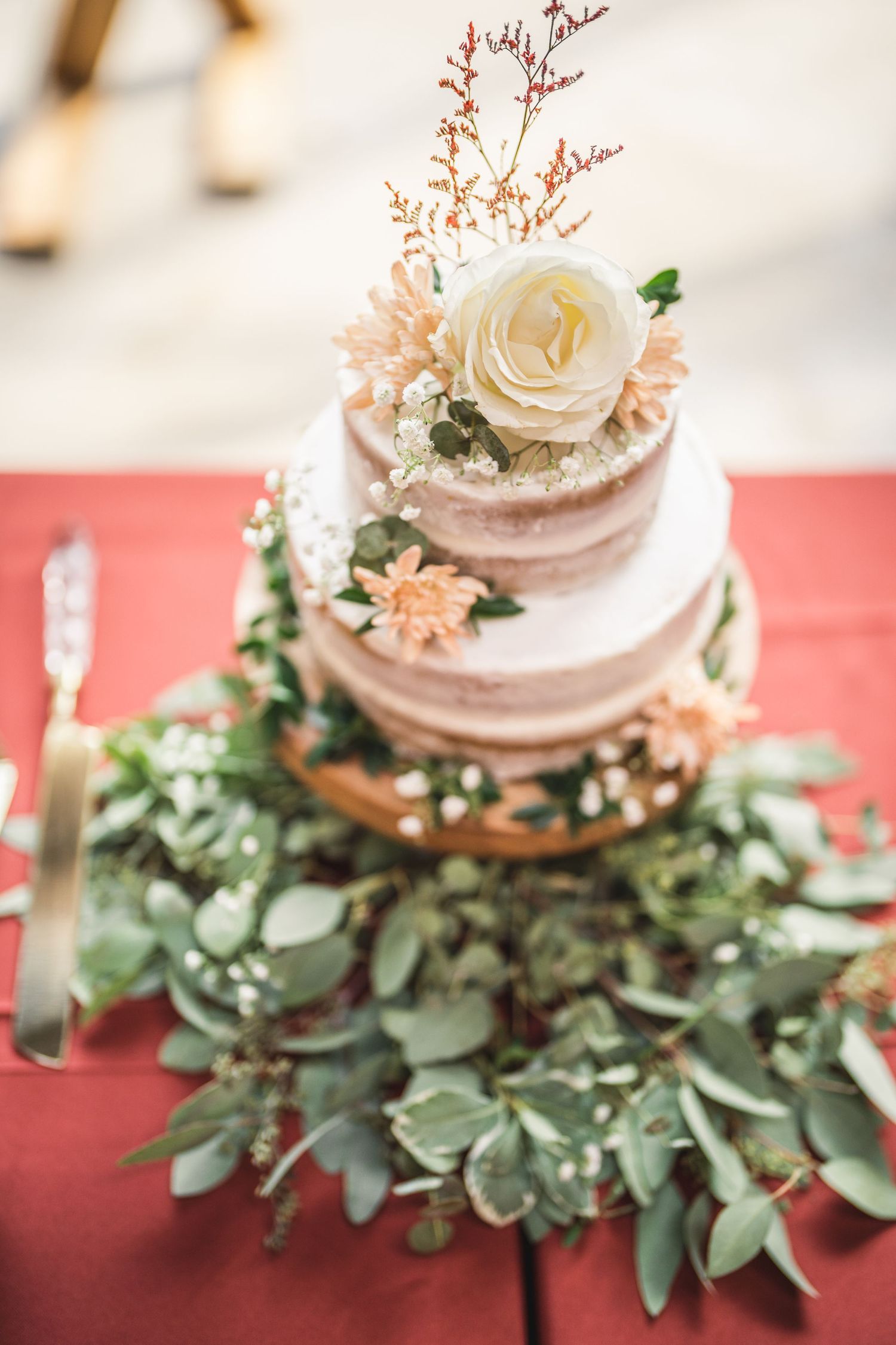 Elegant wedding cake with white rose and greenery decorations on a red tablecloth.