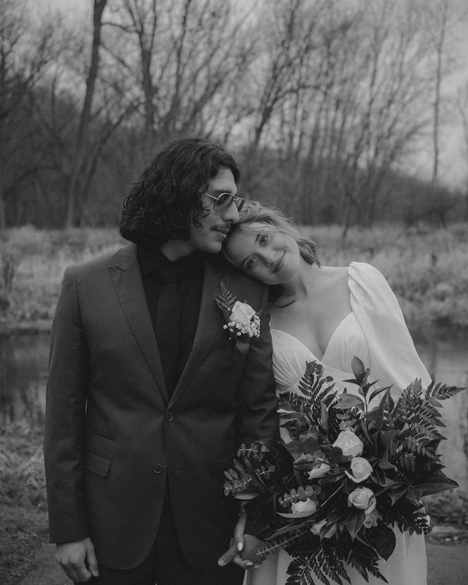A romantic black and white wedding portrait of a couple by a lake with the bride holding a lush bouquet.