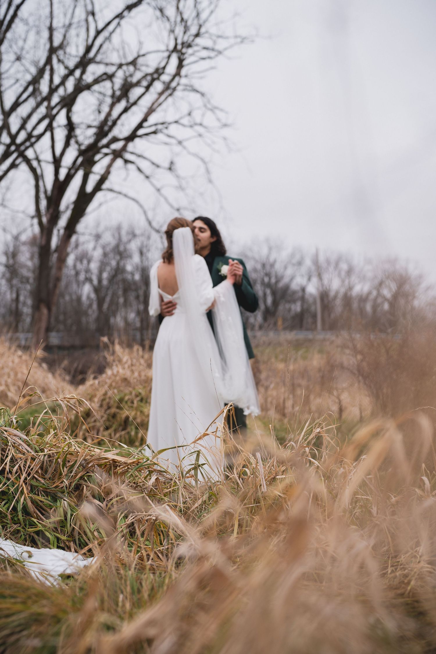 A couple in wedding attire embraces in a winter field with bare trees and dried grass in the background.