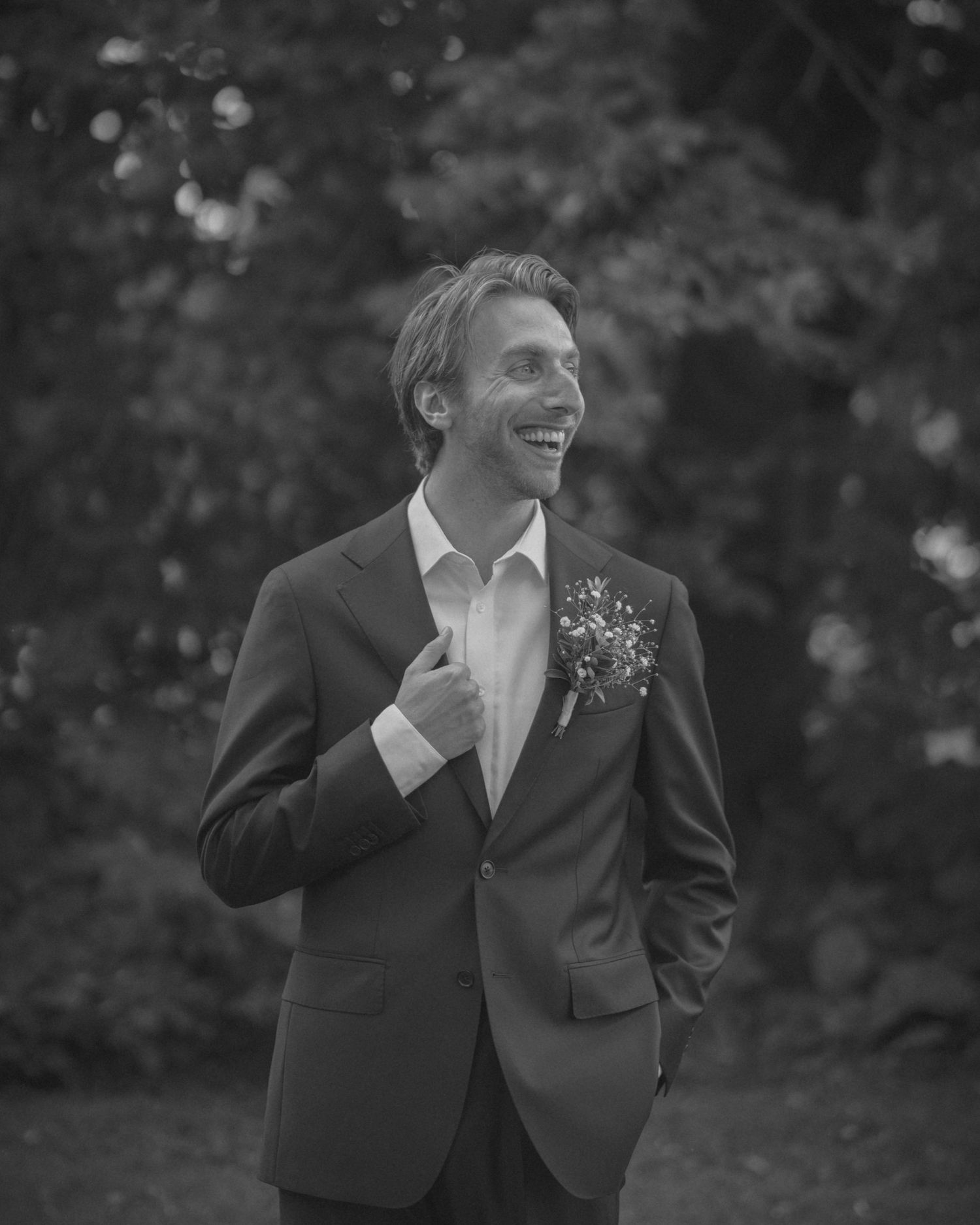 Black and white portrait of smiling groom in dark suit with boutonniere among green trees.