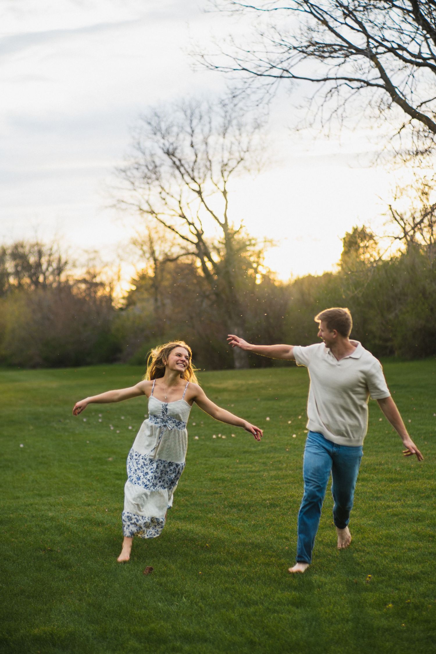 A laughing couple runs barefoot across a lush green lawn at sunset with trees in the background.