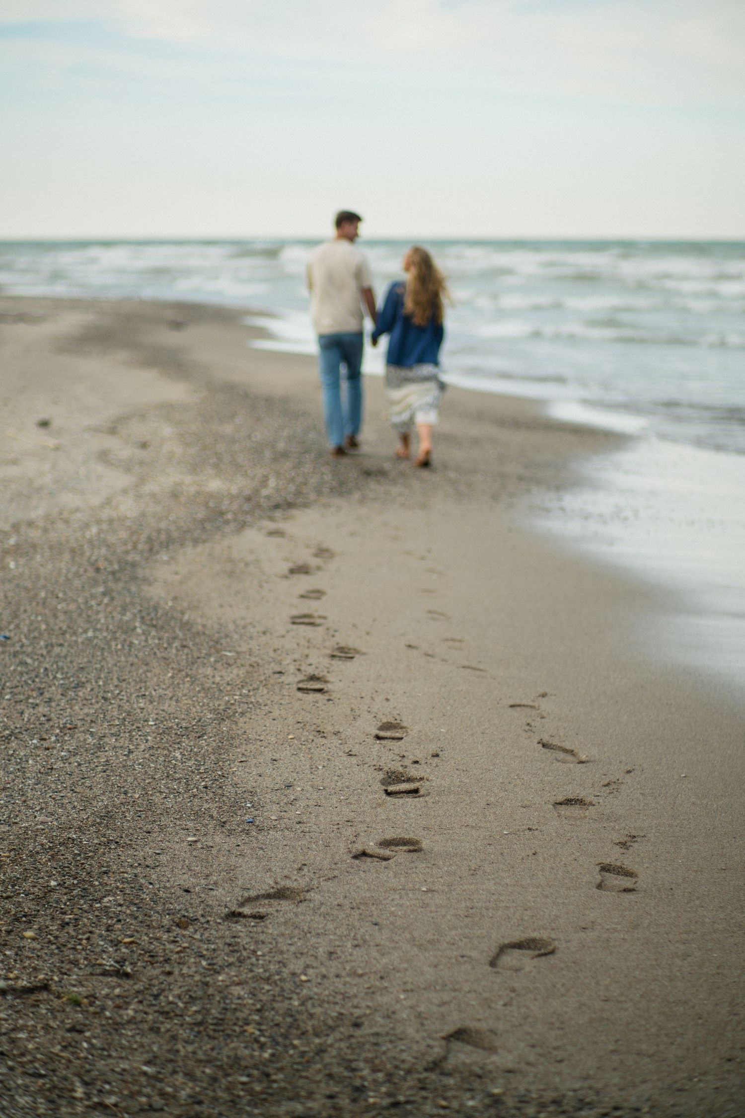 Couple holding hands walking along sandy beach leaving footprints in the sand with ocean waves in background.