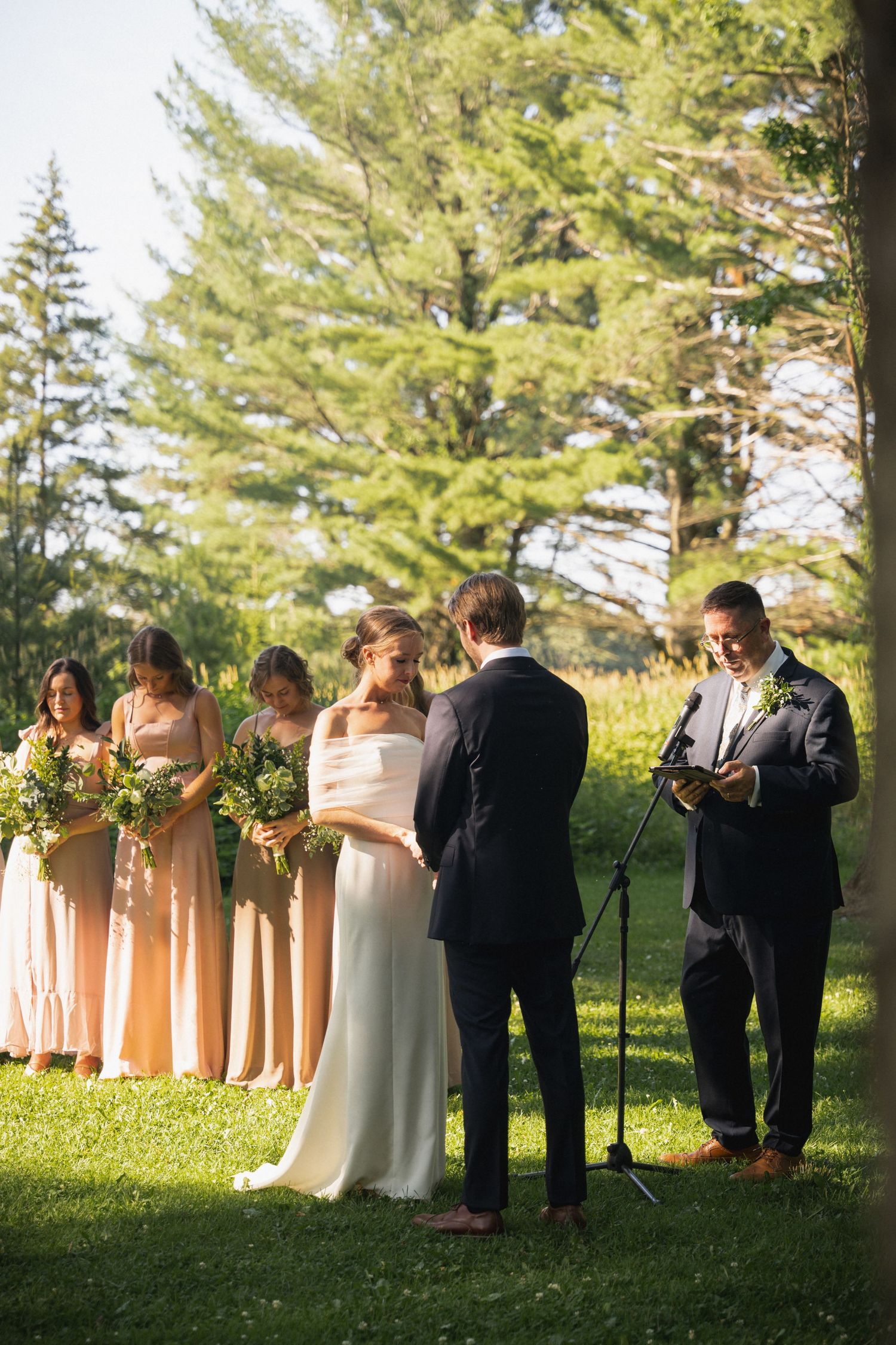A bride and groom exchange vows outdoors at sunset with bridesmaids in peach dresses standing nearby.