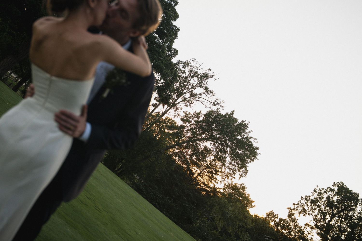 Bride and groom share a romantic kiss at sunset on a lush green lawn.