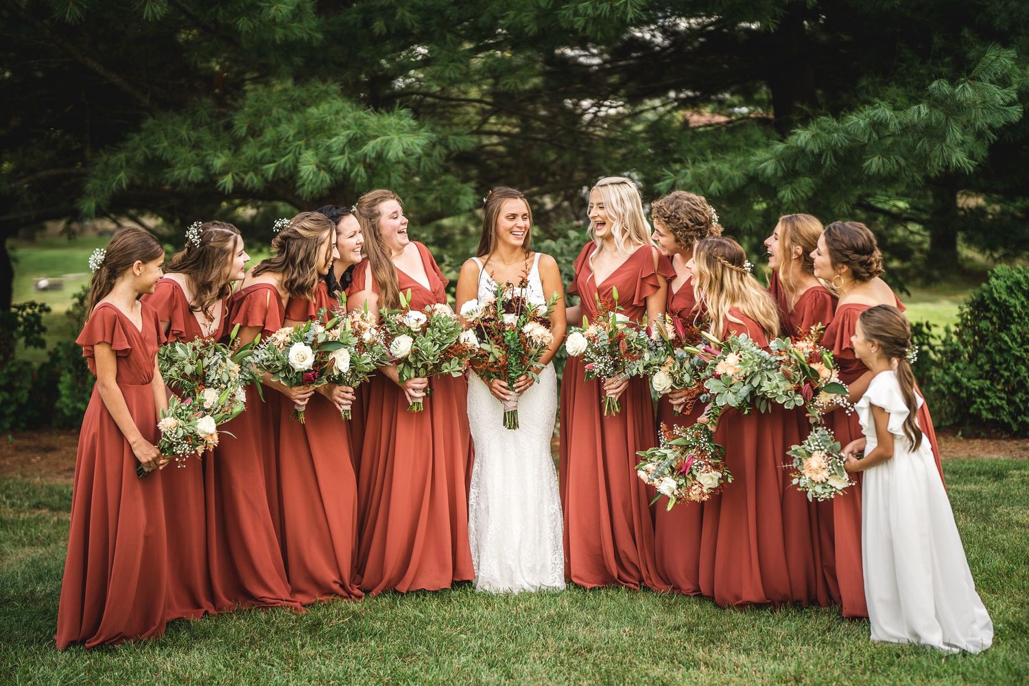 Formal wedding party portrait with bridesmaids in rust gowns on green lawn.