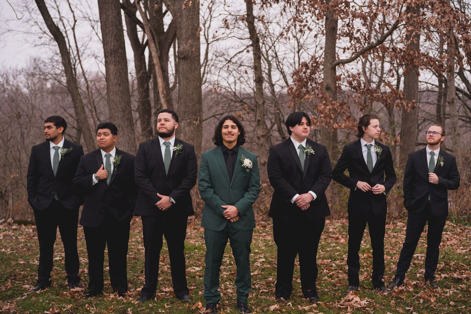 Wedding party groomsmen line up in black suits with one wearing emerald green in wooded setting.