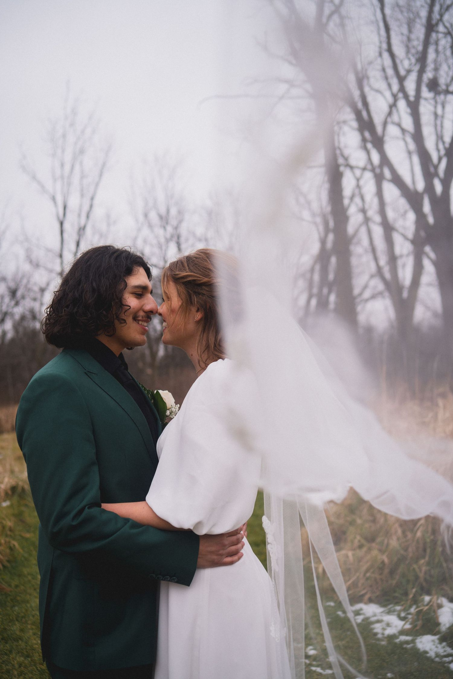 A couple shares an intimate moment in wedding attire with a flowing veil in a winter landscape.