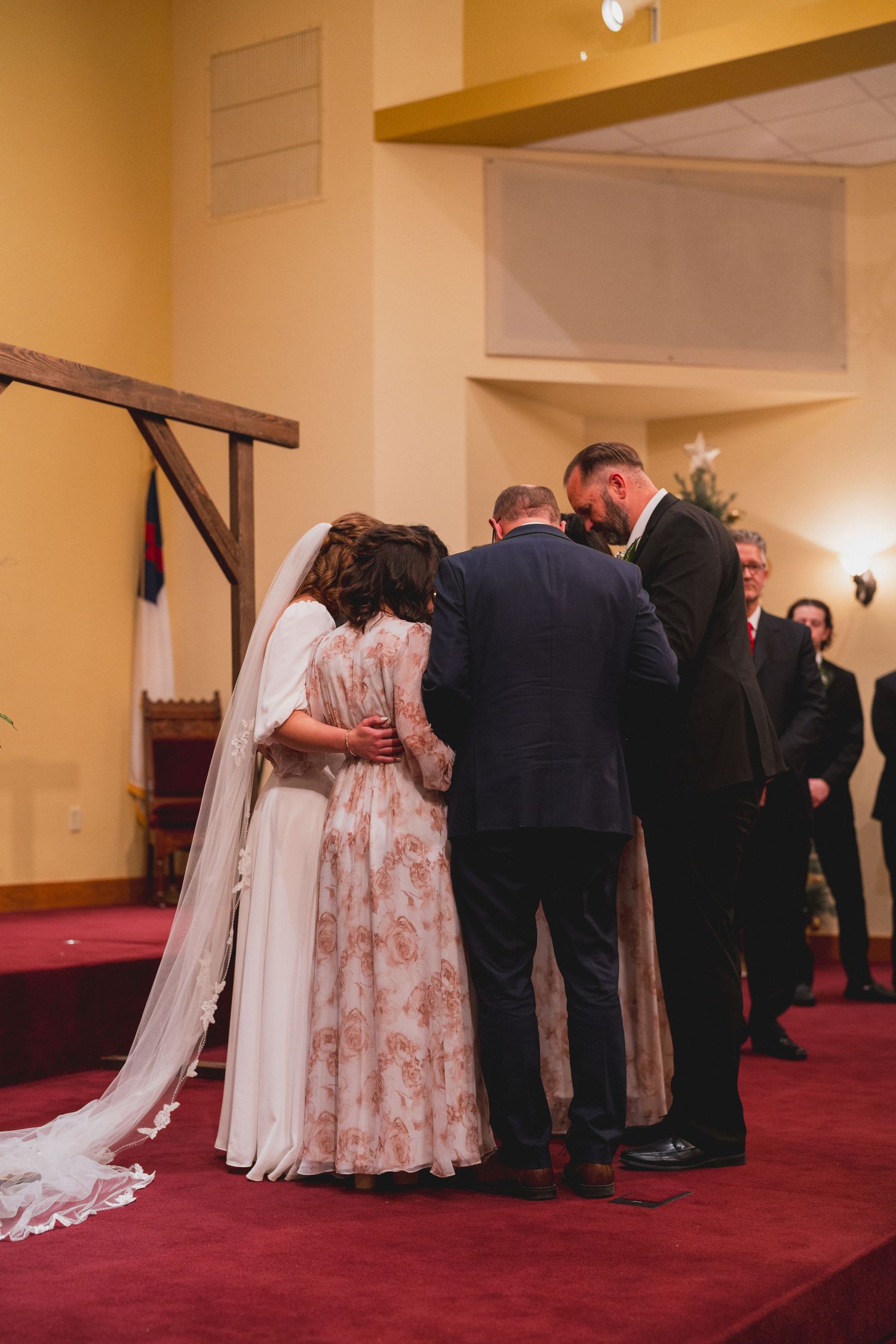 Wedding party gathers in prayer during ceremony on red carpeted church stage.