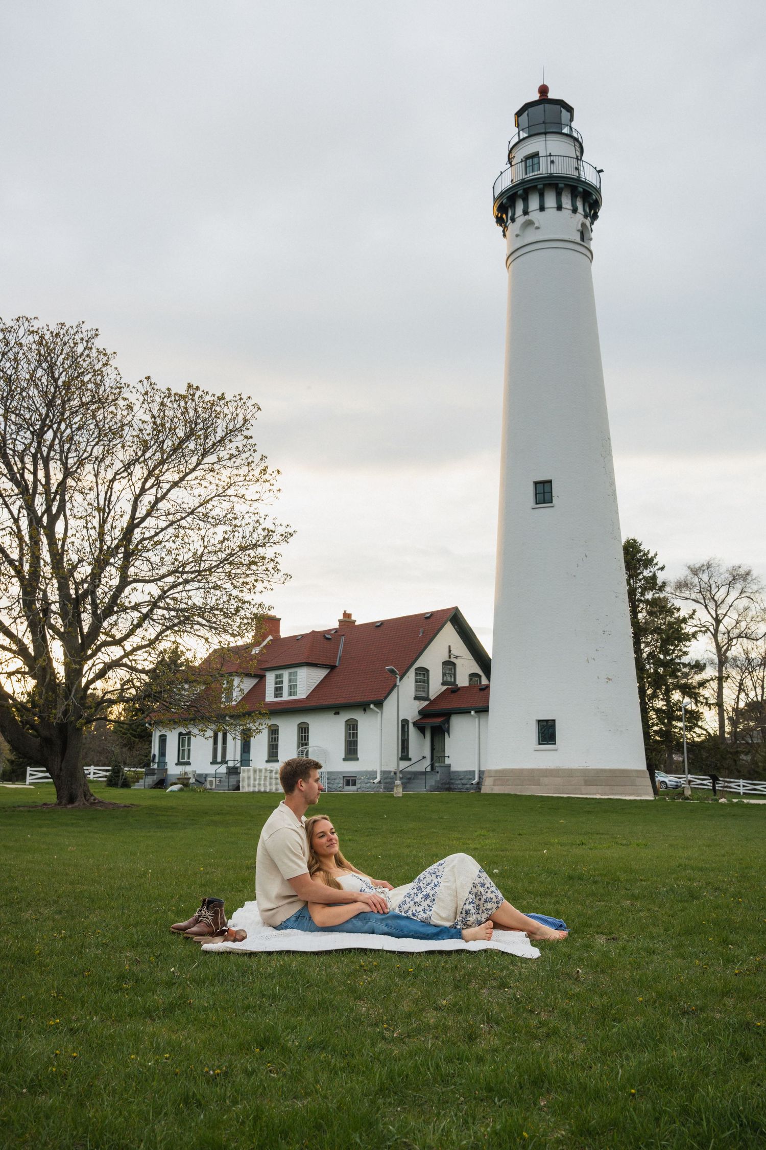 Couple relaxing on a blanket on green grass in front of a tall white lighthouse.
