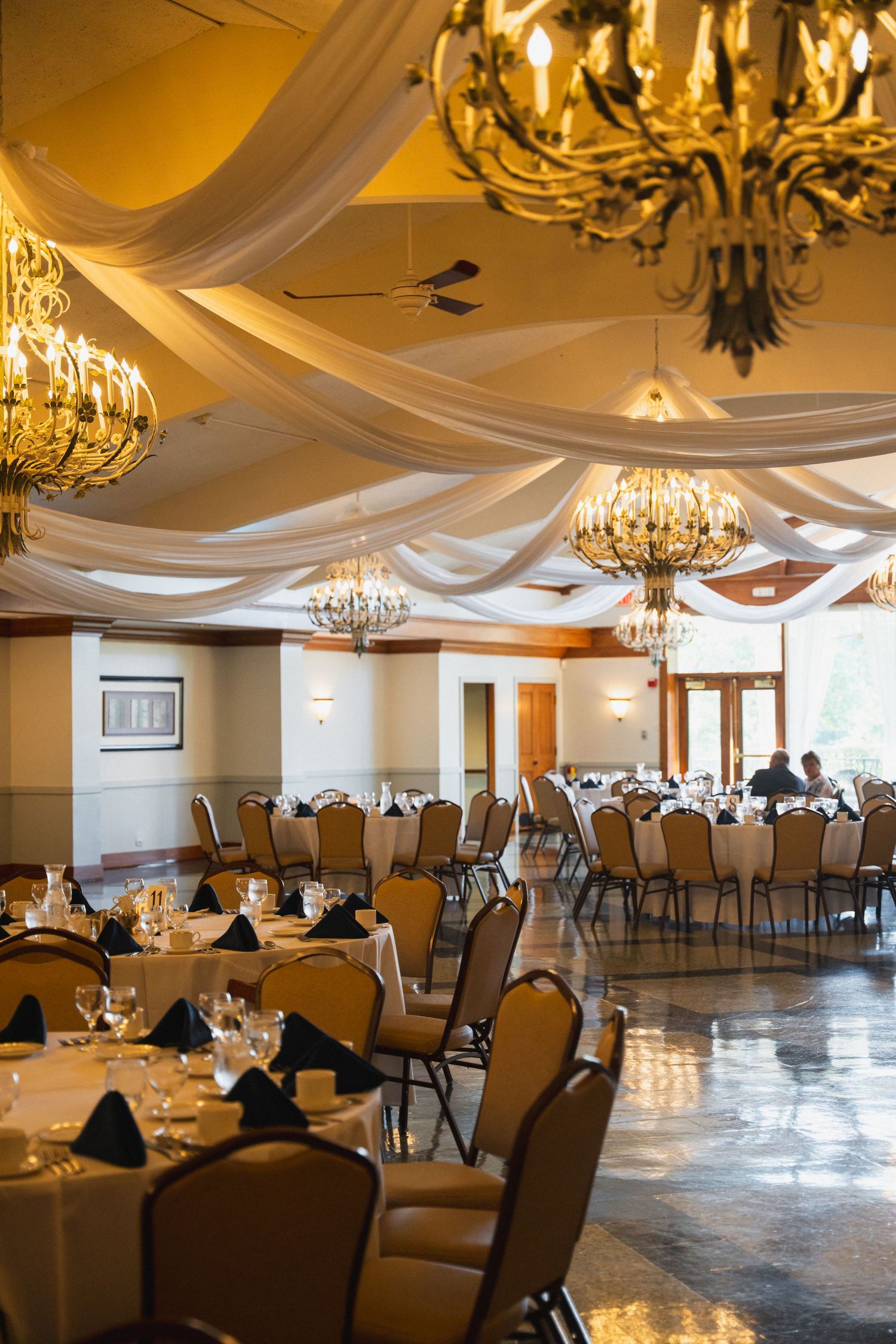 Elegant wedding reception ballroom with crystal chandeliers, draped white fabric ceiling, and round tables.