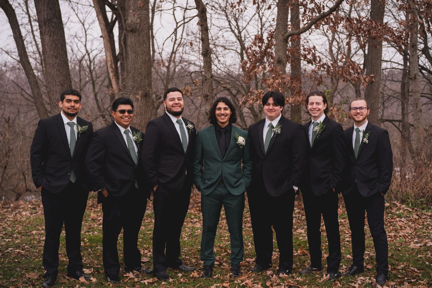 Groomsmen in black suits with one member in emerald green pose in autumn forest setting.
