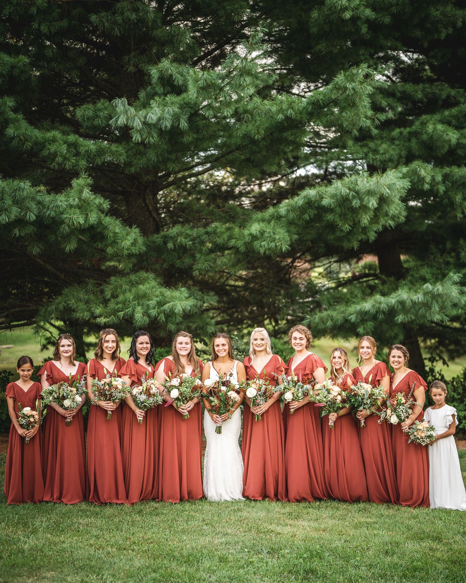 Wedding party photo with bridesmaids in rust-colored dresses against pine tree backdrop.