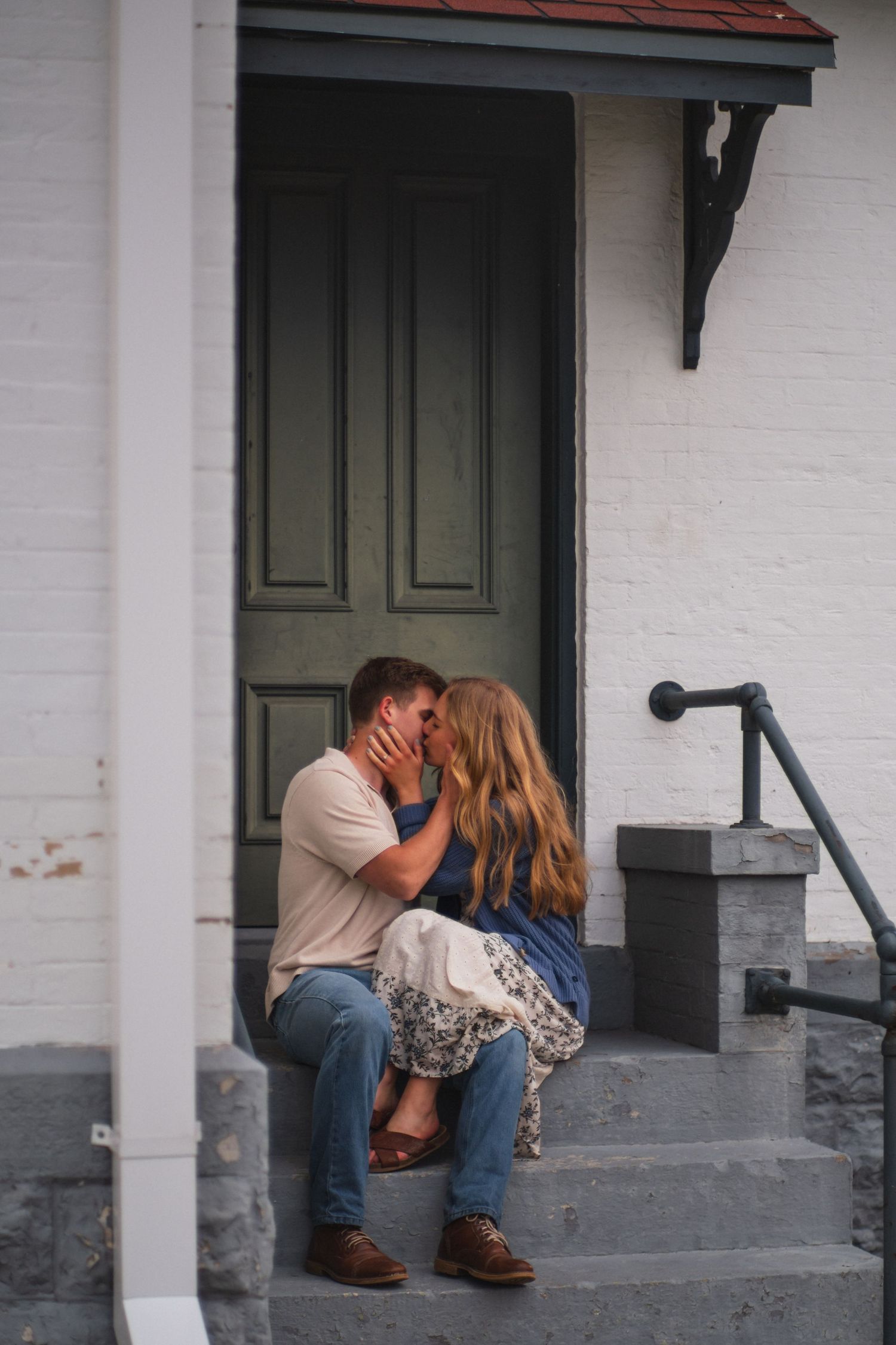 Couple sharing a kiss while seated on steps of a white brick building with a dark green door.