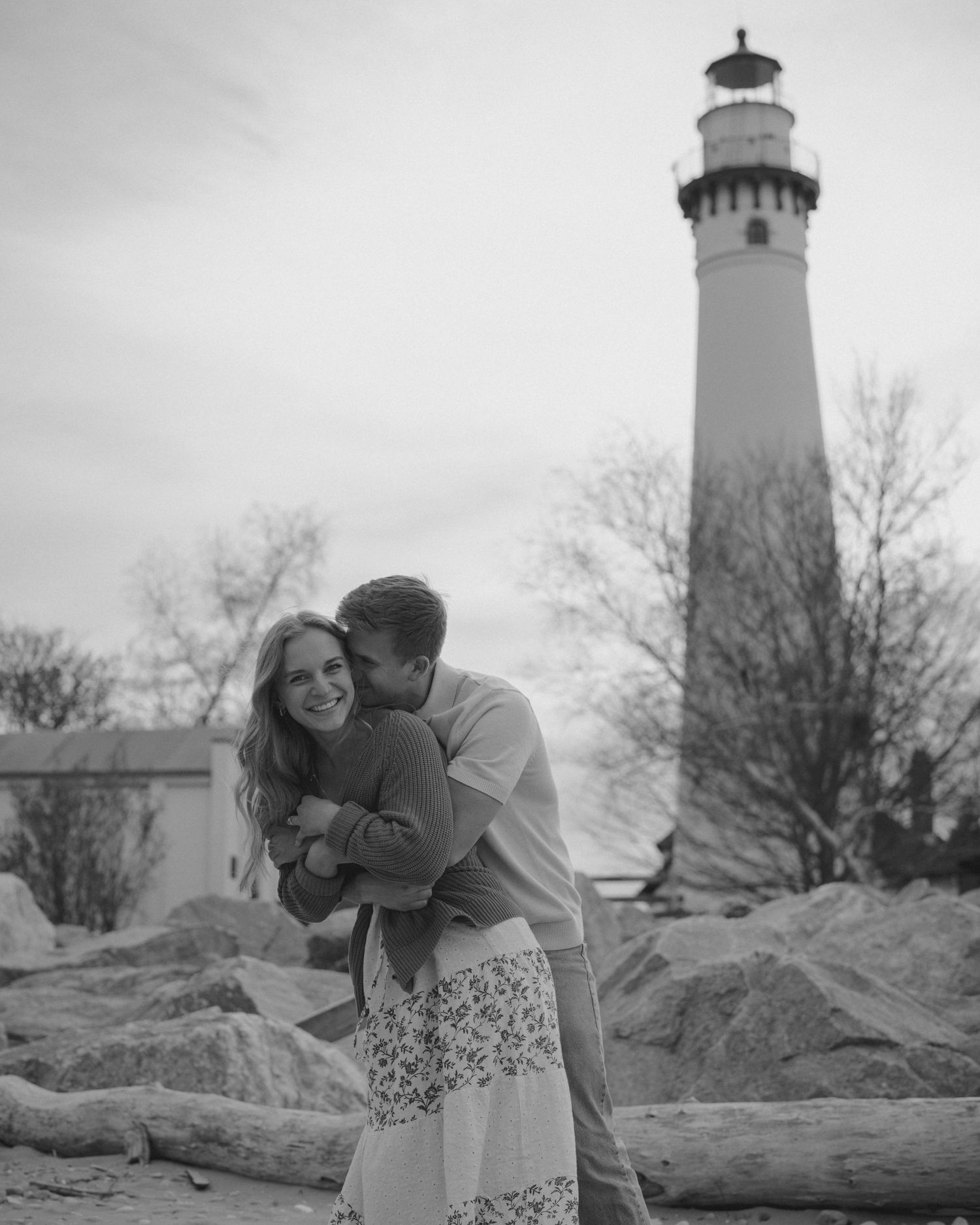 Black and white photo of a couple embracing near a lighthouse on a rocky beach.