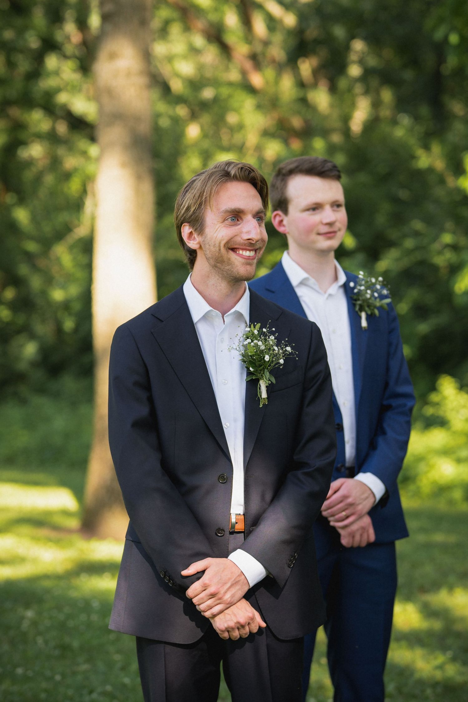 Smiling groom in dark suit with boutonniere standing outdoors with groomsman in navy suit behind him.