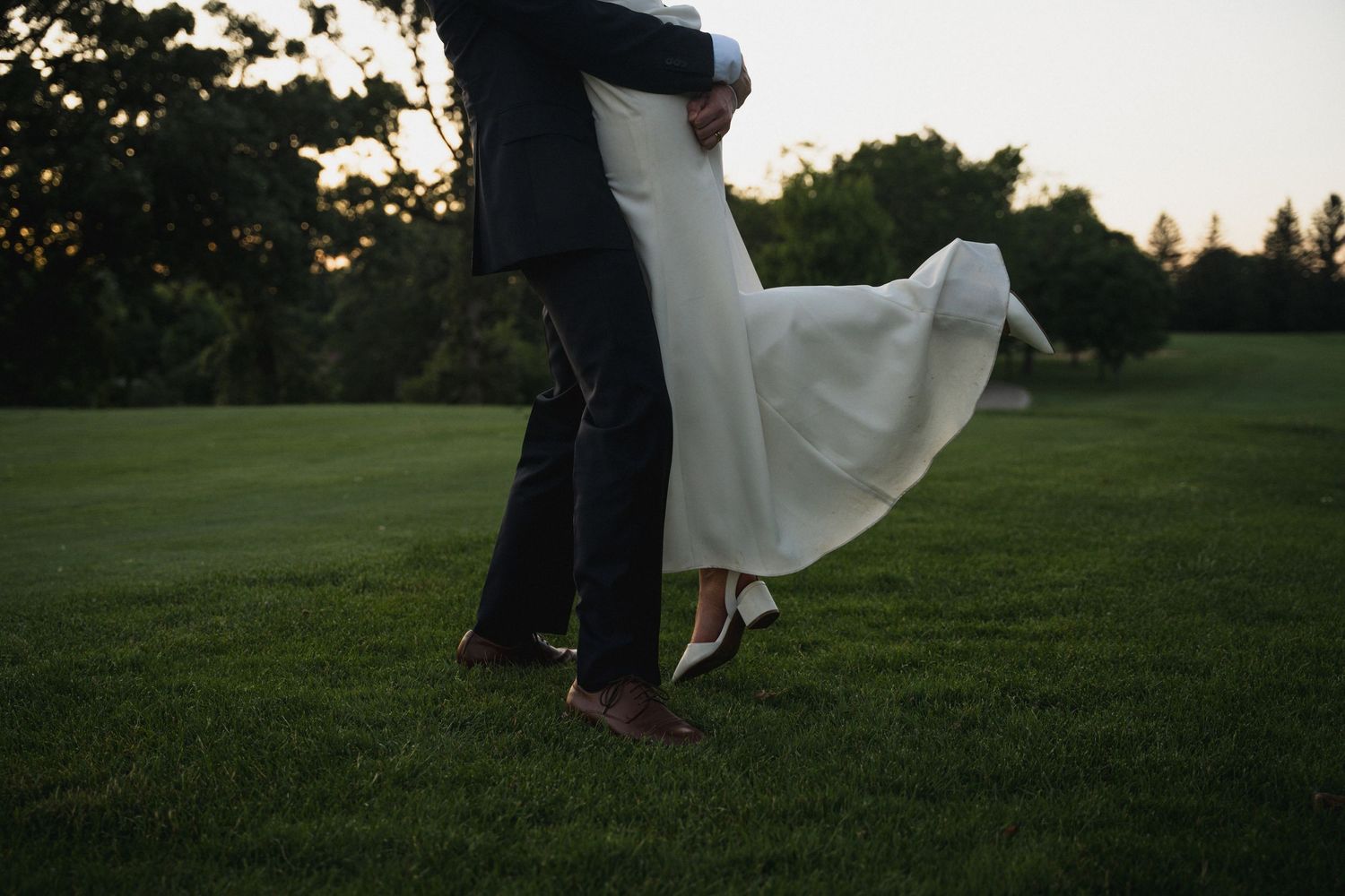 Groom lifting bride in flowing white wedding dress on green lawn at golden hour sunset.