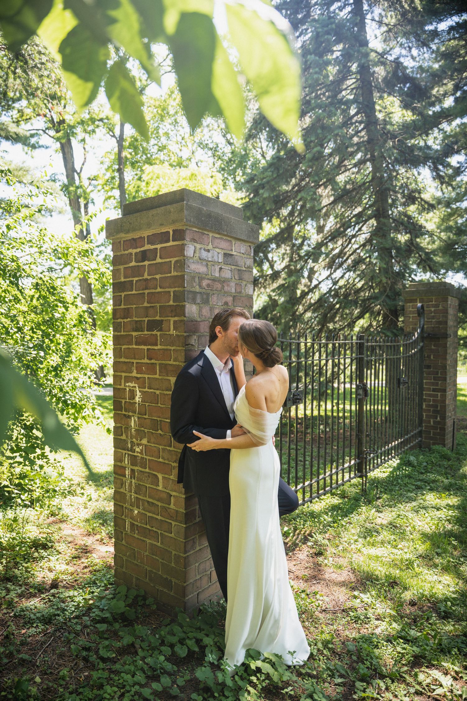 A bride and groom kiss beside a brick gate pillar surrounded by lush green trees outdoors.