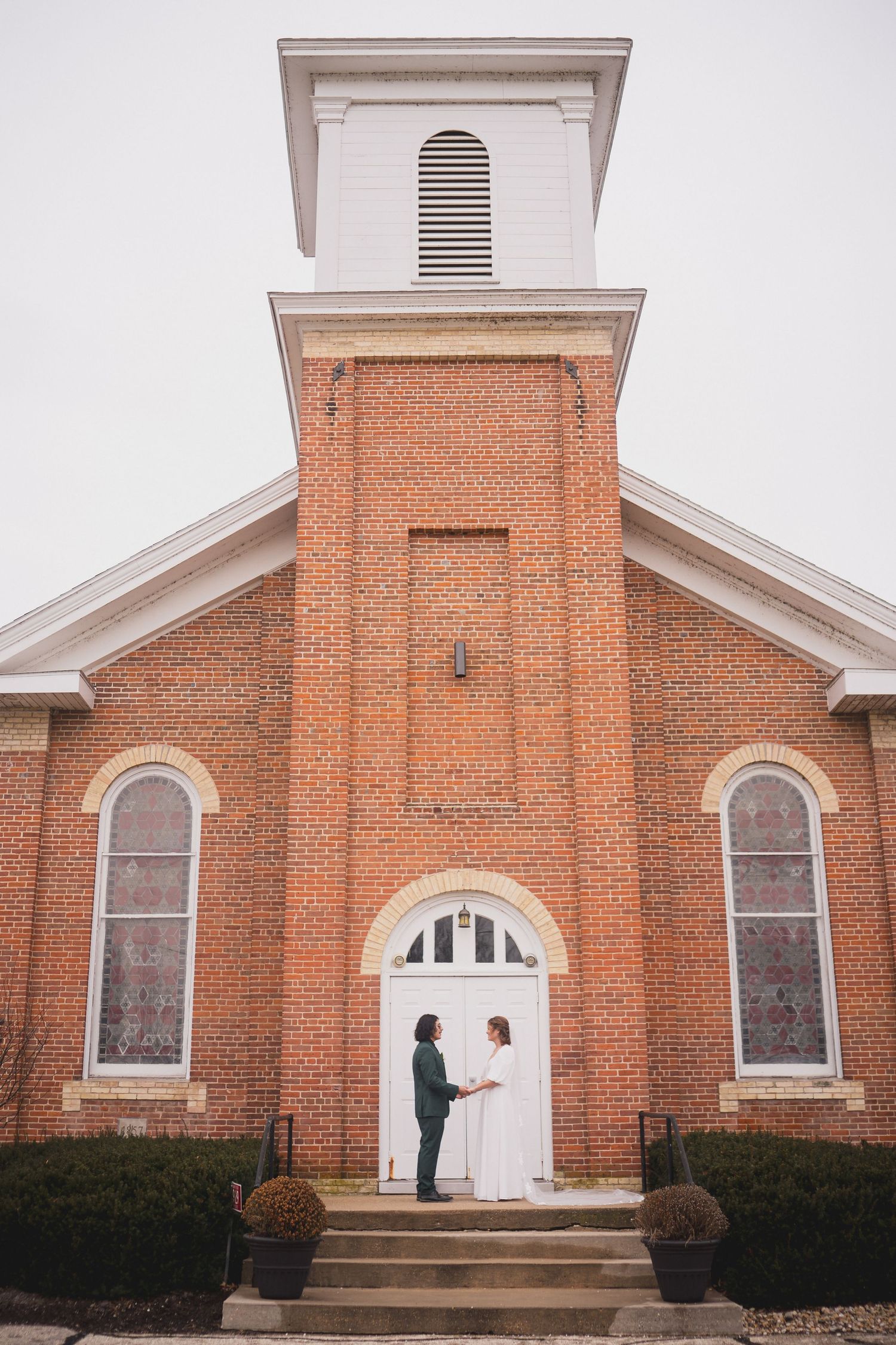Wide view of a red brick church exterior with a couple standing by the entrance.