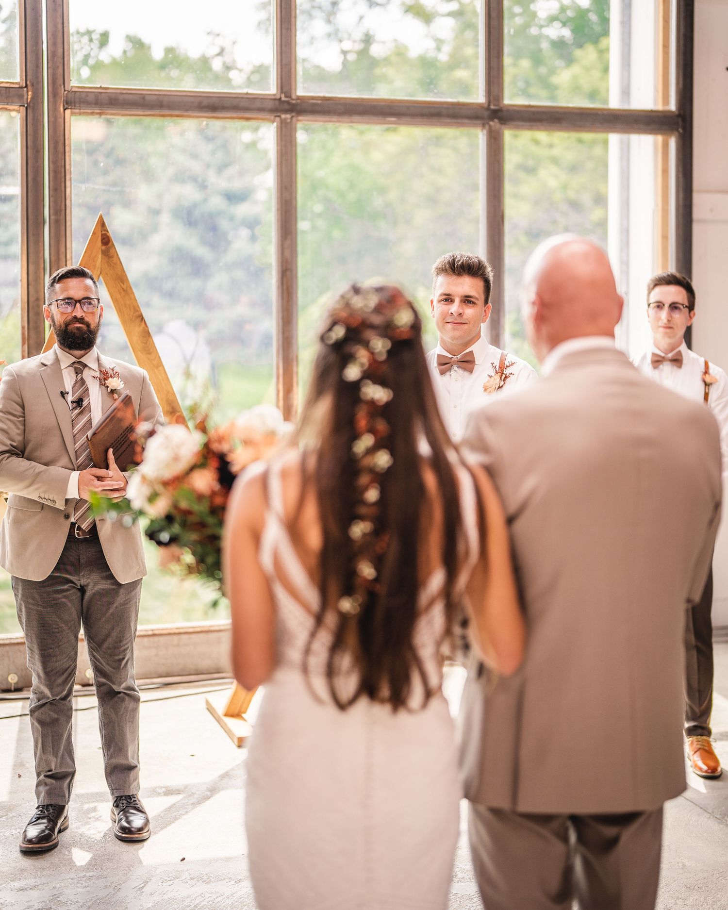 Wedding ceremony in progress with geometric wooden backdrop and natural light streaming through large windows.