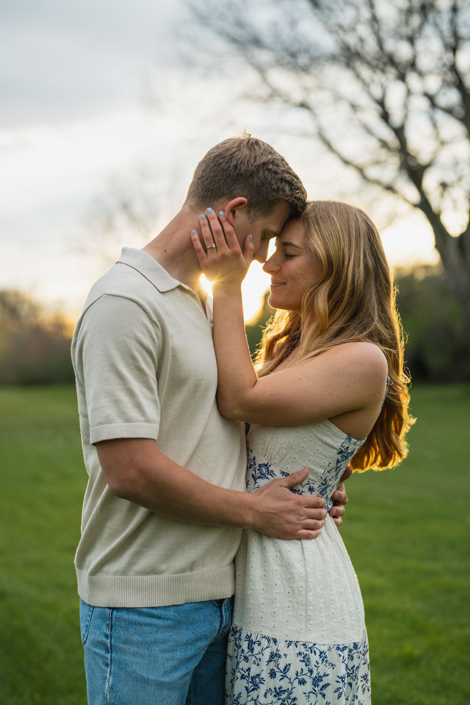Romantic couple touching foreheads at golden sunset in green field, woman in floral dress.