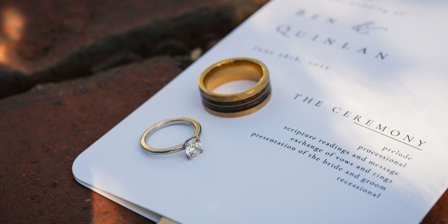 Gold wedding band and diamond engagement ring resting on a wedding ceremony program for Ben and Quinlan.