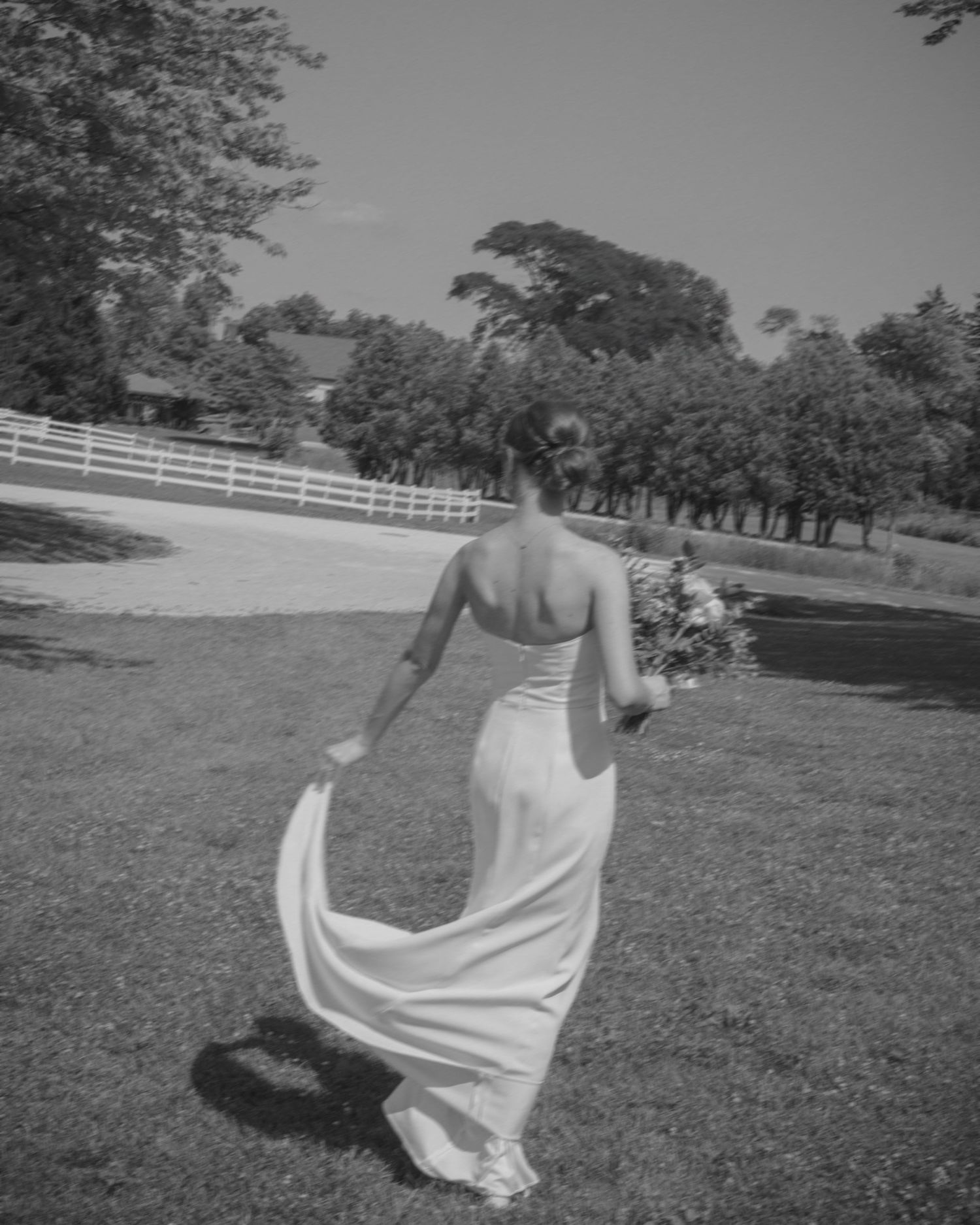Black and white photo of bride in backless wedding gown walking along white fence holding bridal bouquet.