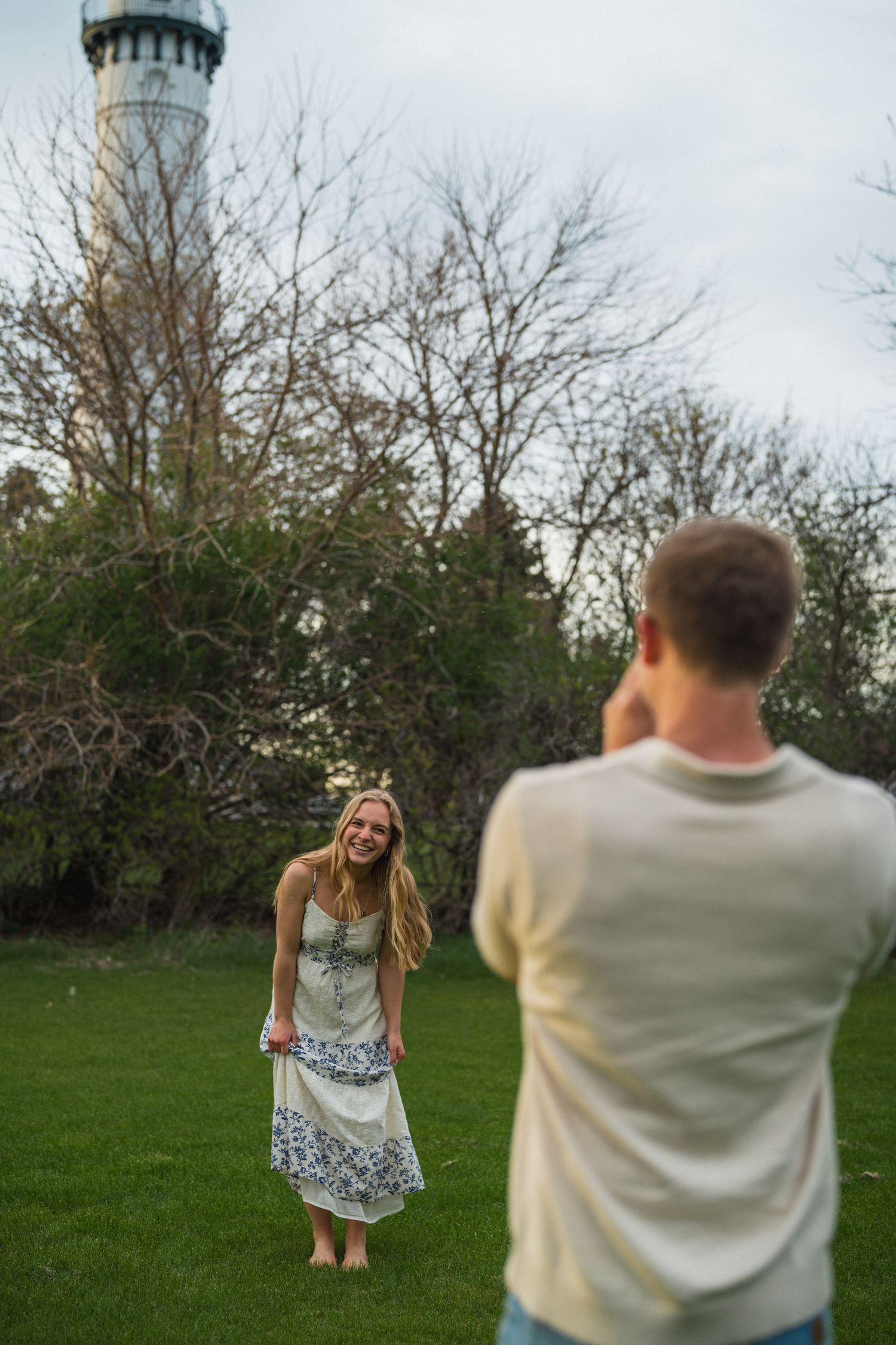 Man photographing smiling blonde woman in floral dress on green lawn near tall lighthouse tower.
