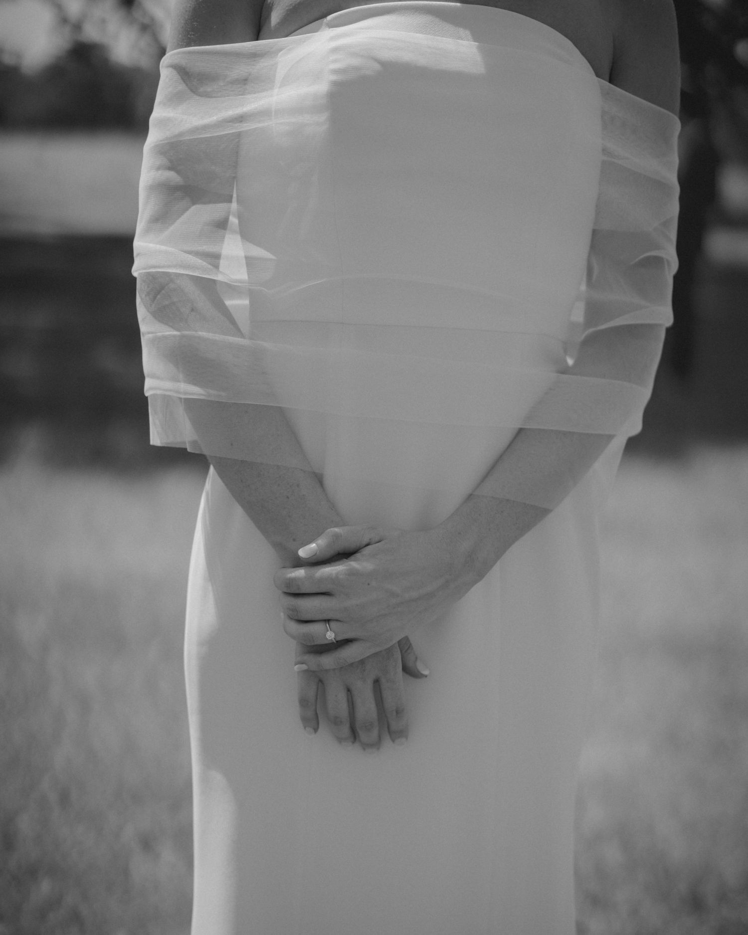 Black and white close-up of bride in strapless wedding gown with tulle detail, hands clasped at waist.