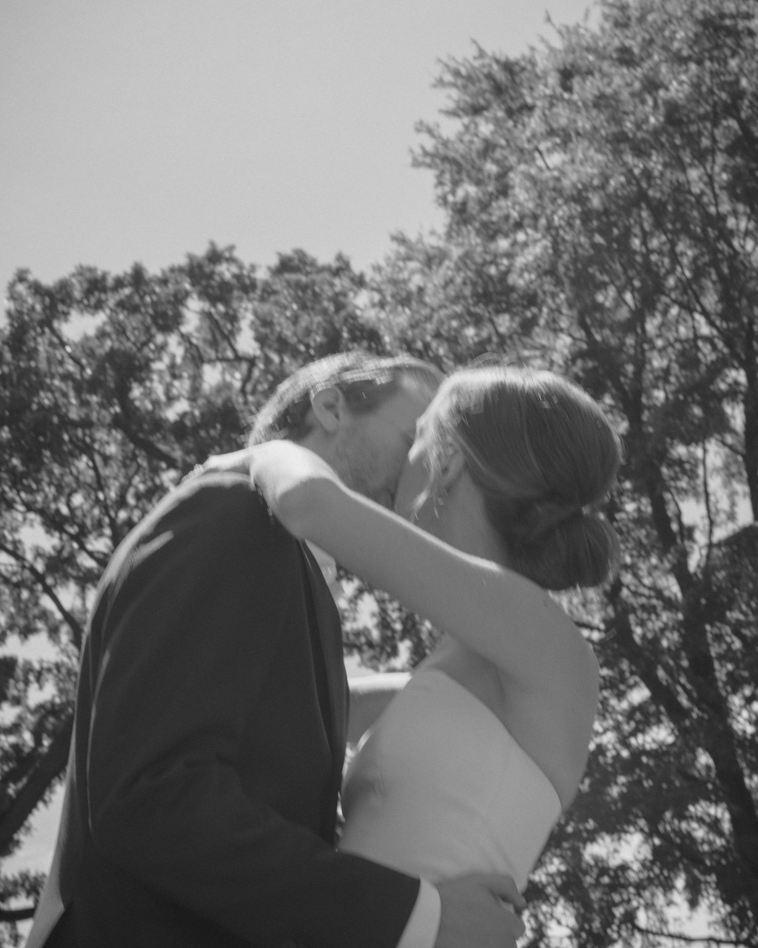 Black and white photo of bride and groom sharing a first kiss outdoors with trees in background.