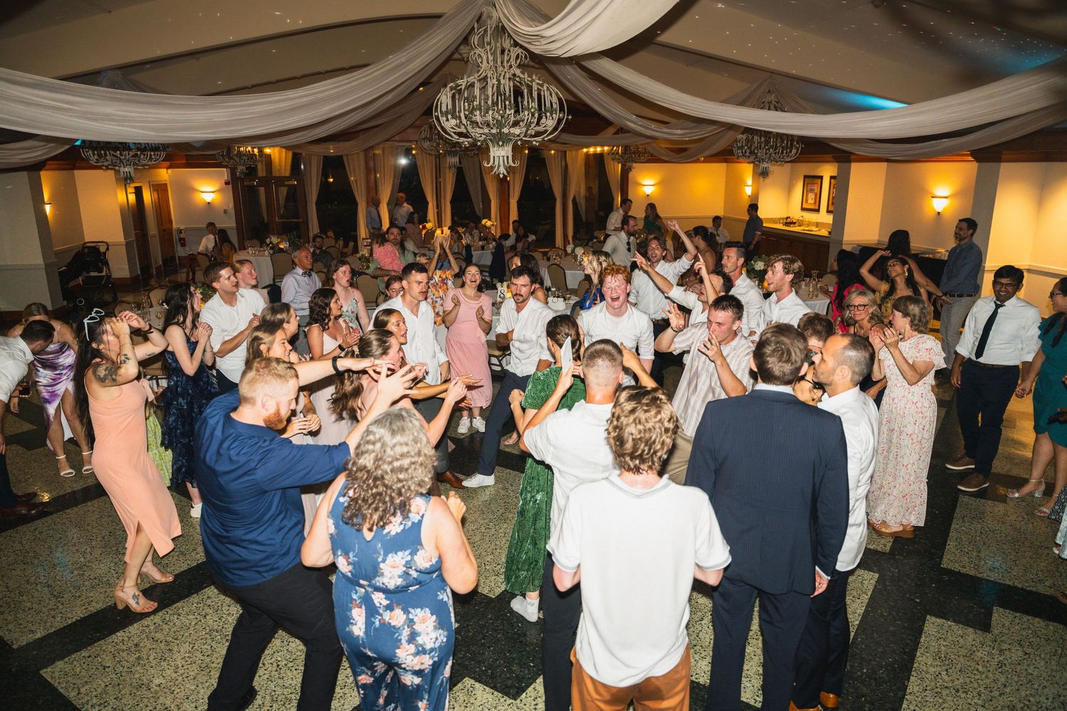 Energetic wedding reception dance floor packed with guests celebrating under a chandelier in a ballroom.