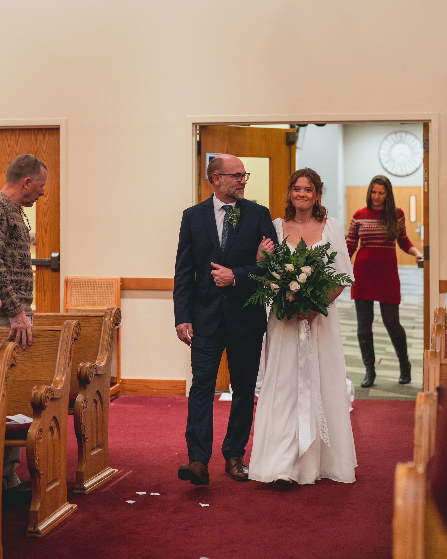 Wedding party walking down red-carpeted church aisle with wooden pews.