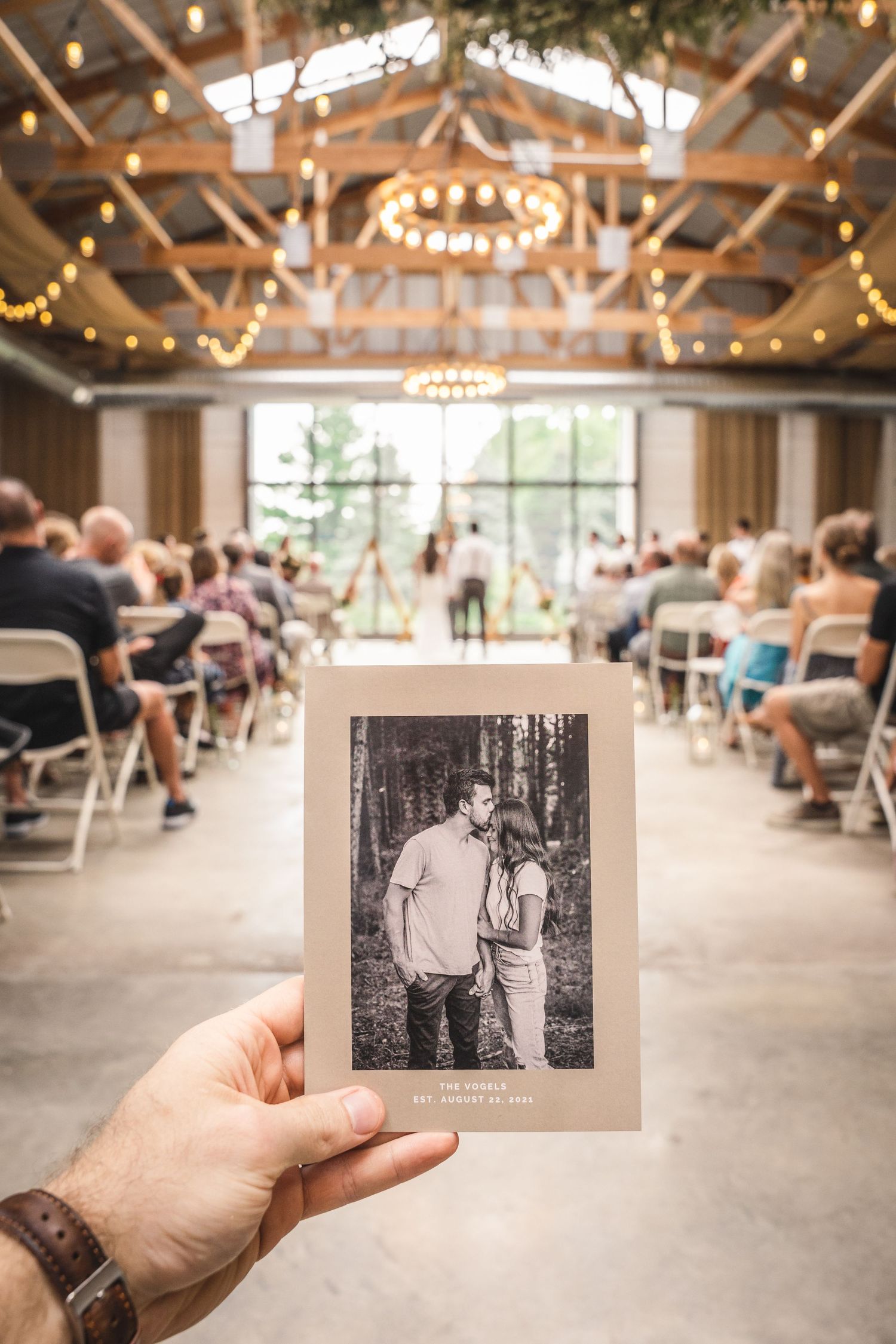 Hand holds black and white photo in foreground of wedding ceremony venue with string lights and wooden beams.