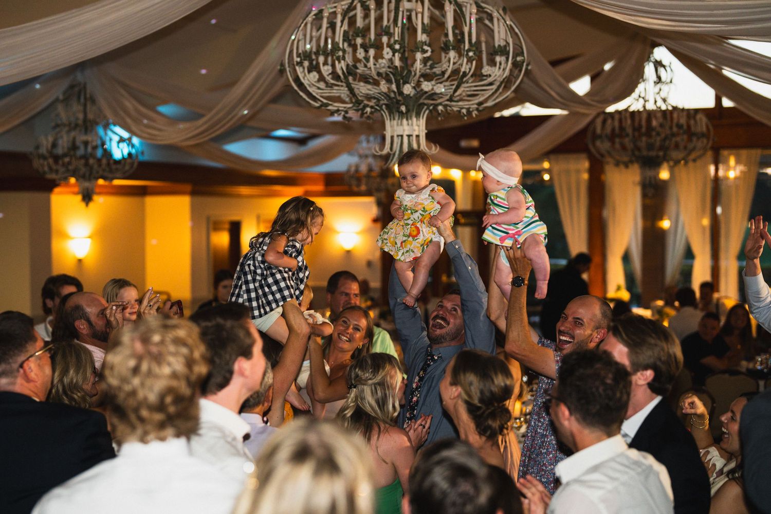 Joyful wedding guests lift babies in the air on a crowded dance floor at reception.