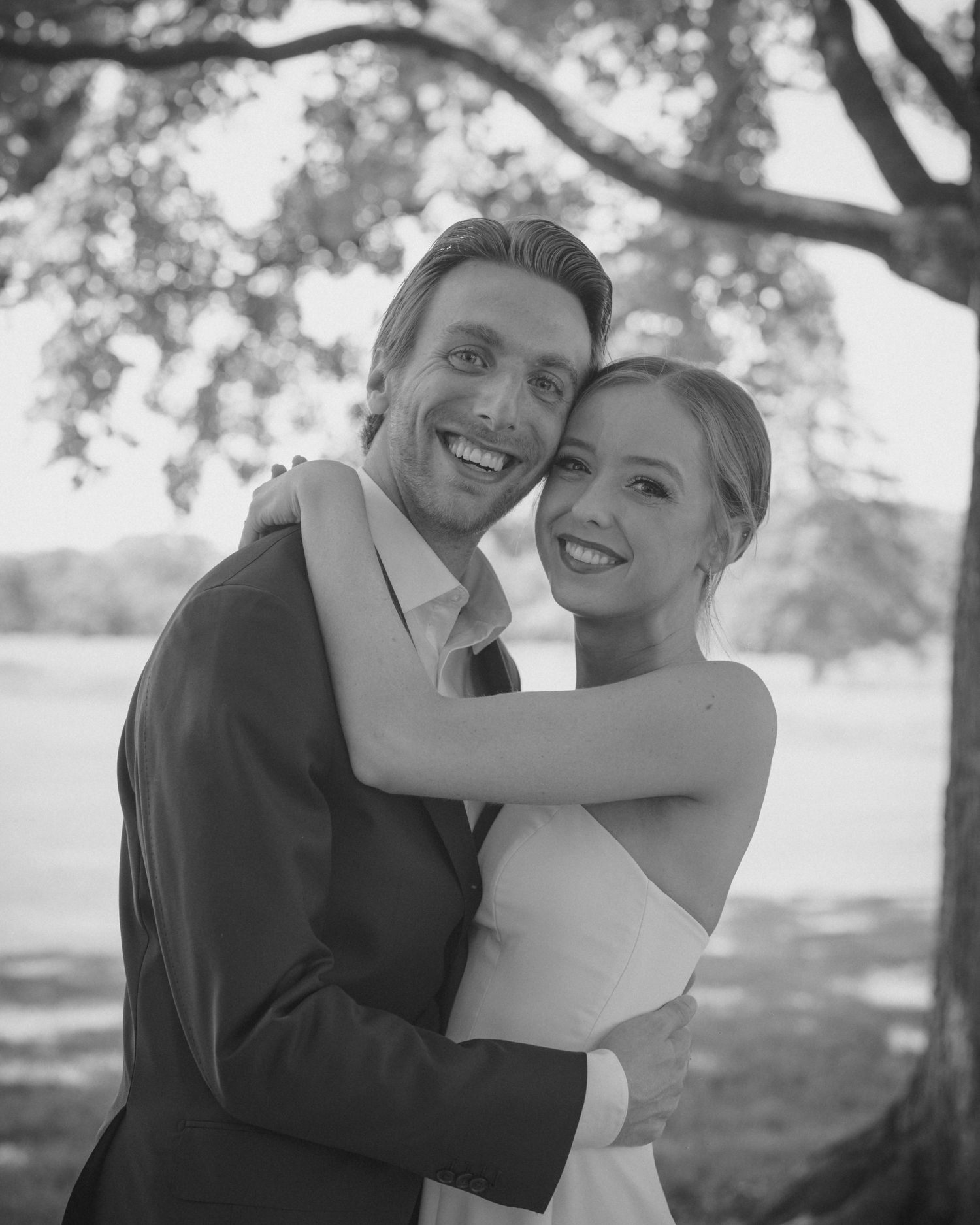 Black and white portrait of smiling bride and groom embracing under a tree on their wedding day.