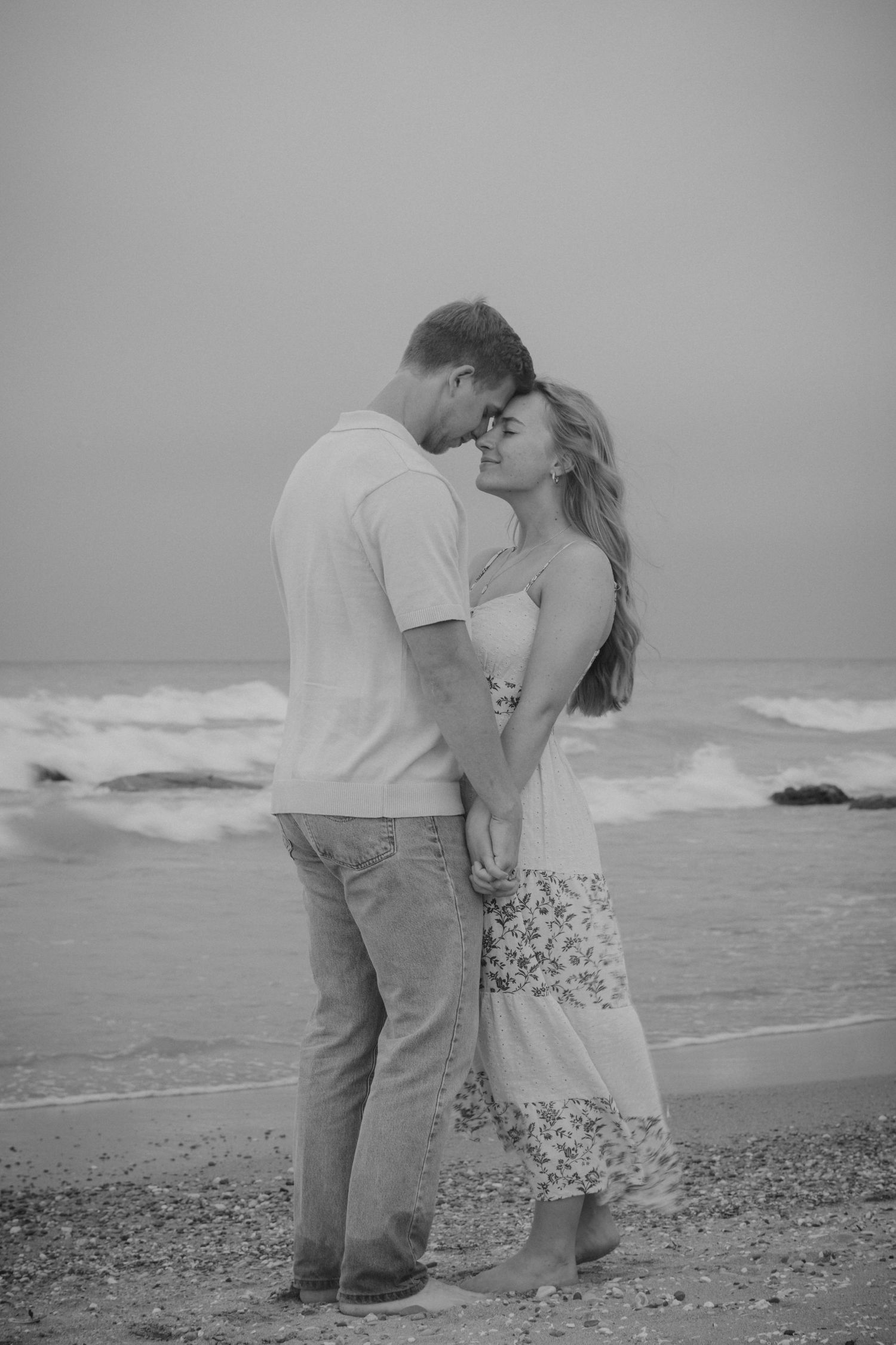 Black and white photo of a romantic couple embracing on a rocky beach with ocean waves in the background.