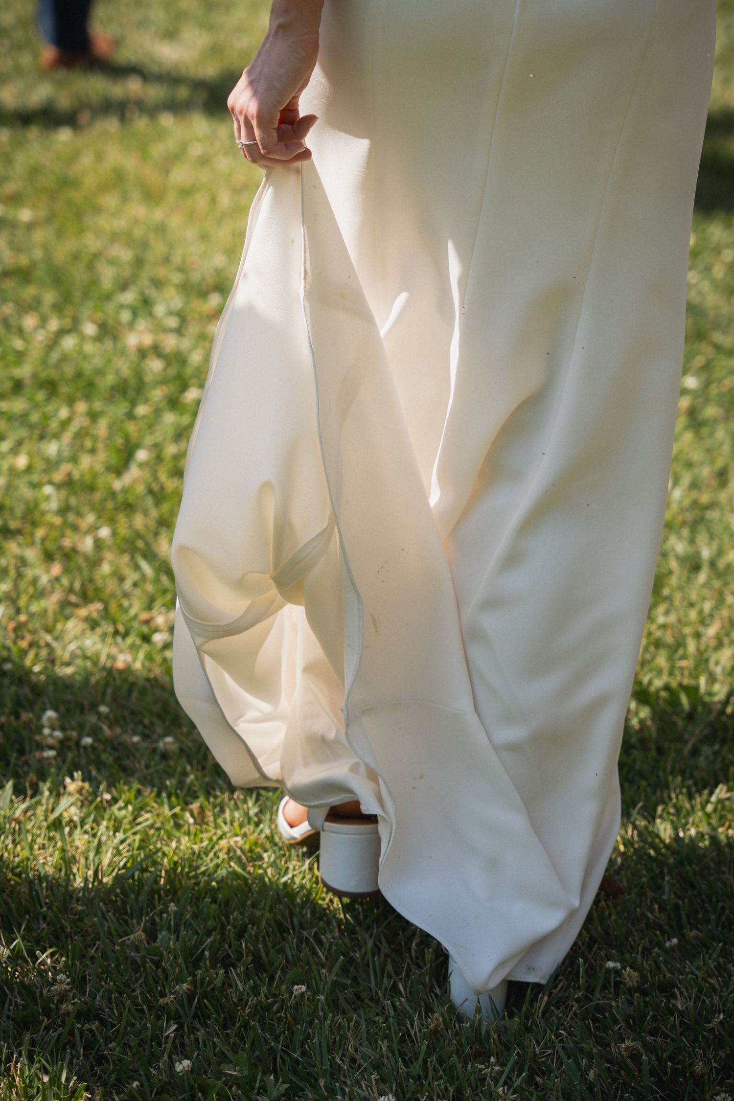 Bride lifting her elegant white wedding gown train while walking on green grass in white heeled sandals.