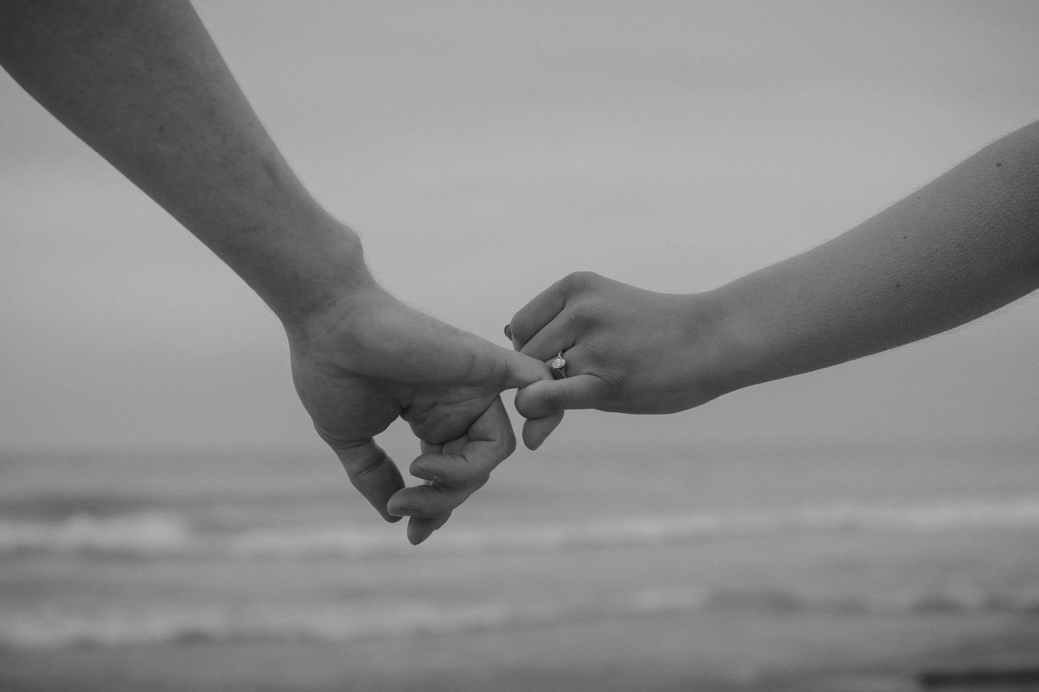 Black and white photo of two people gently holding fingers near ocean beach, romantic couple connection.