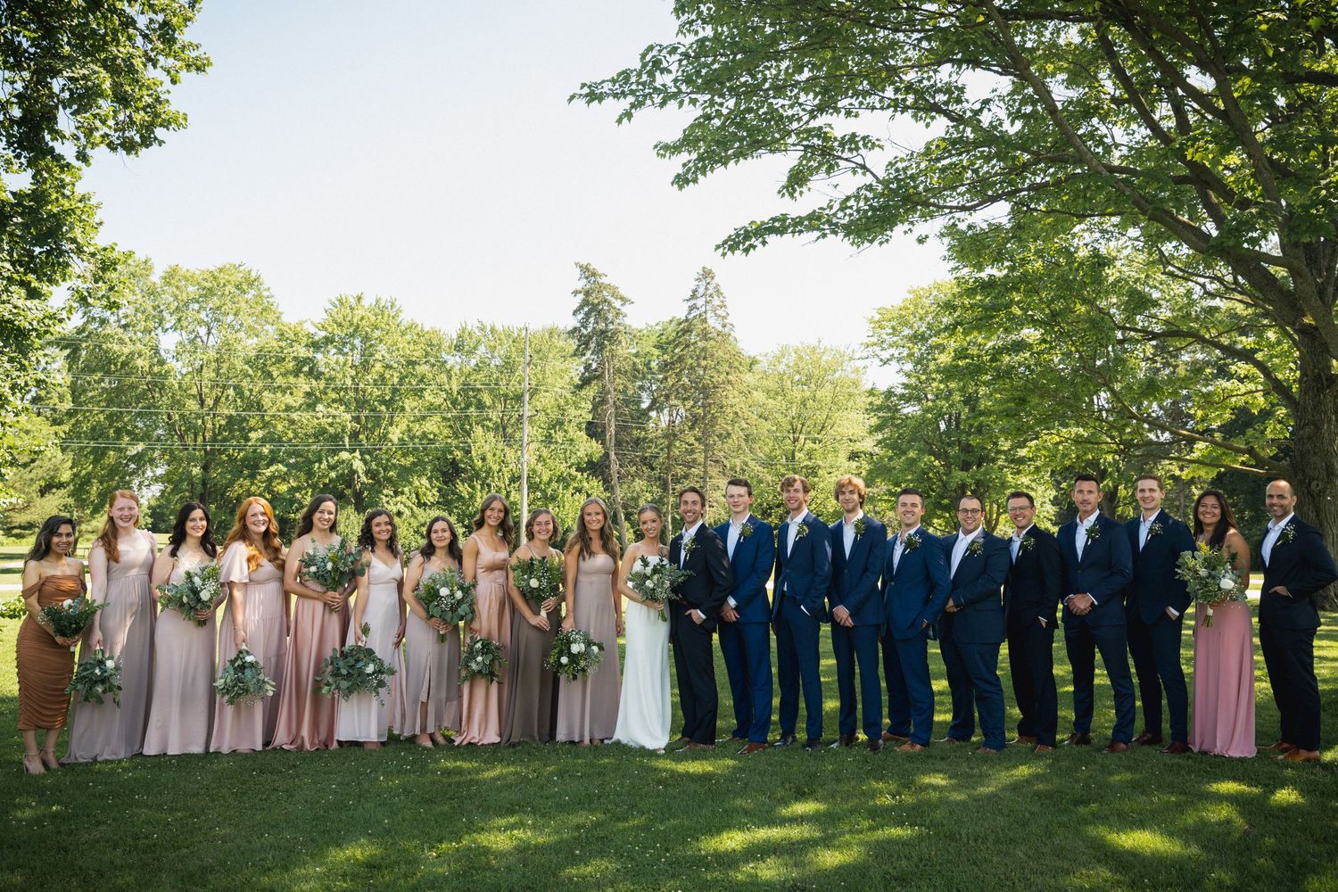 Large wedding party posing outdoors on green lawn with bridesmaids in blush gowns and groomsmen in navy suits.