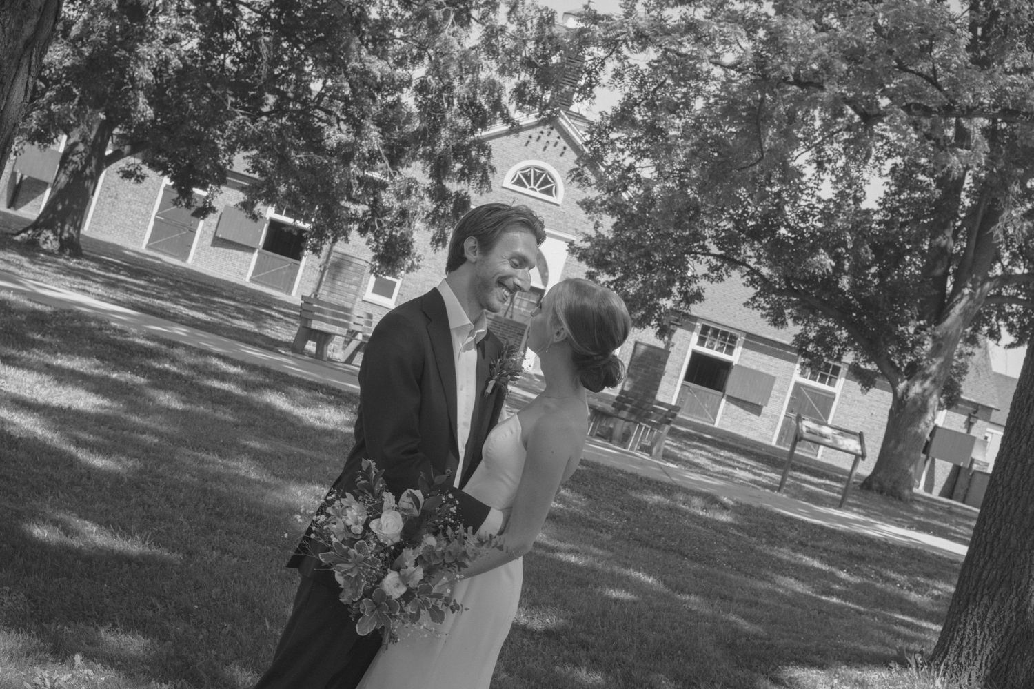 Black and white photo of a bride and groom sharing a romantic moment outdoors under trees near a church.