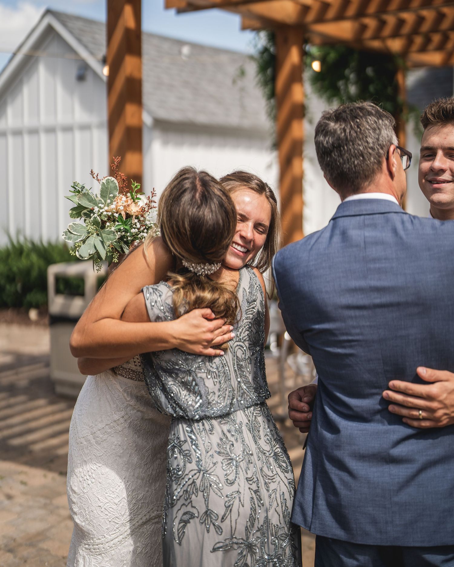 Wedding guests share warm embraces under a wooden pergola.