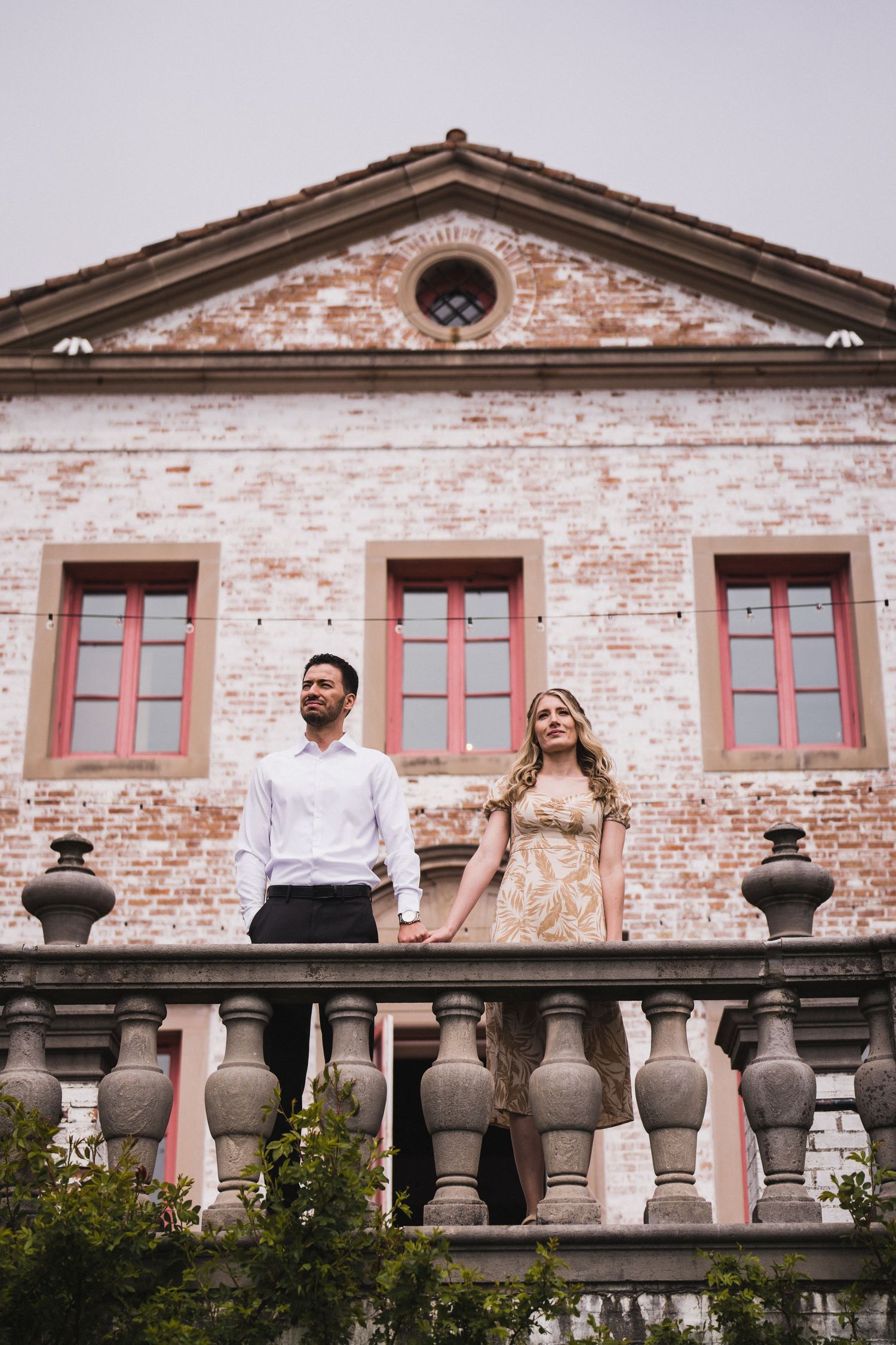 Couple standing behind ornate stone balustrade against weathered brick building with red-trimmed windows.