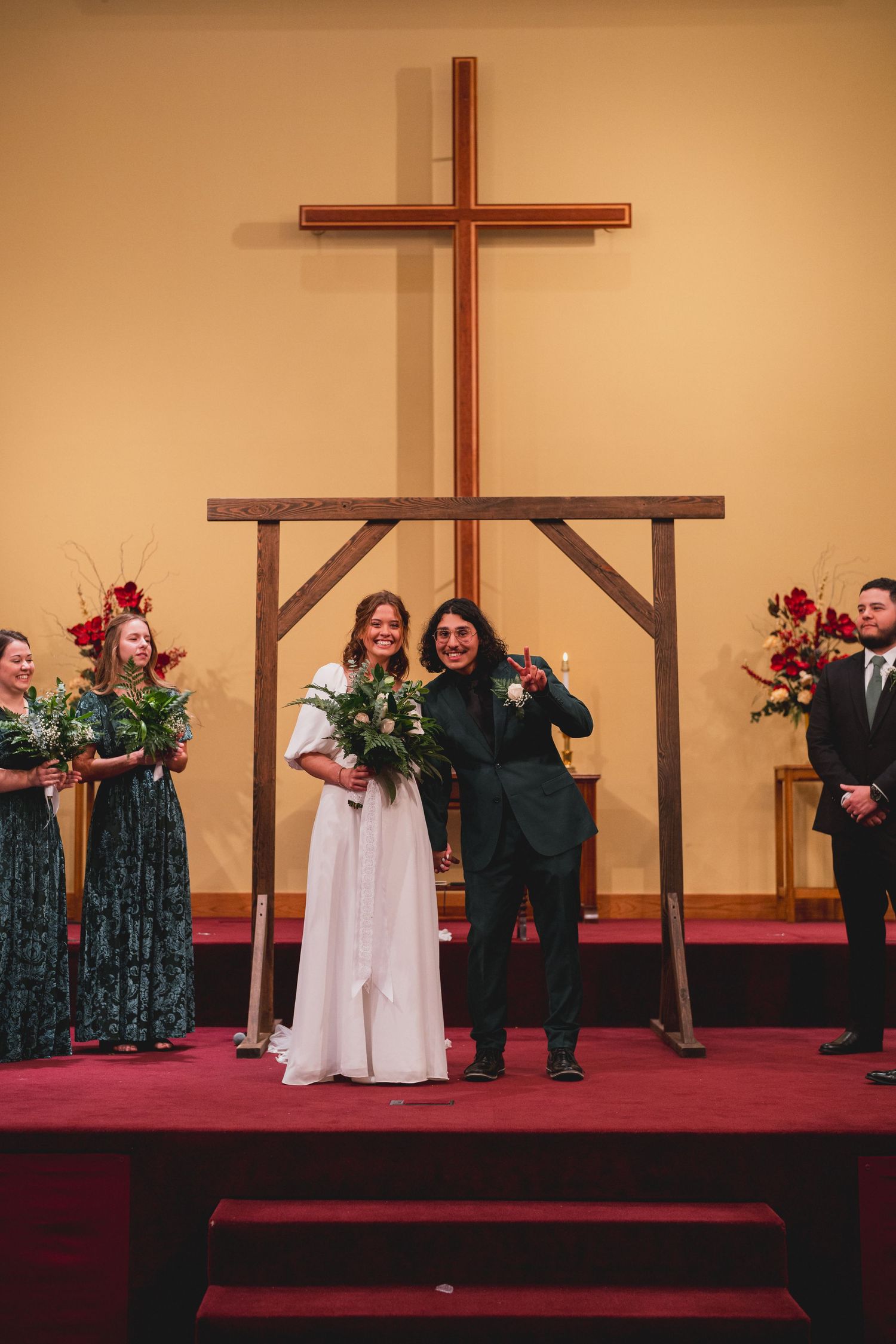 Newlyweds celebrate joyfully under wooden cross with wedding party in emerald dresses.