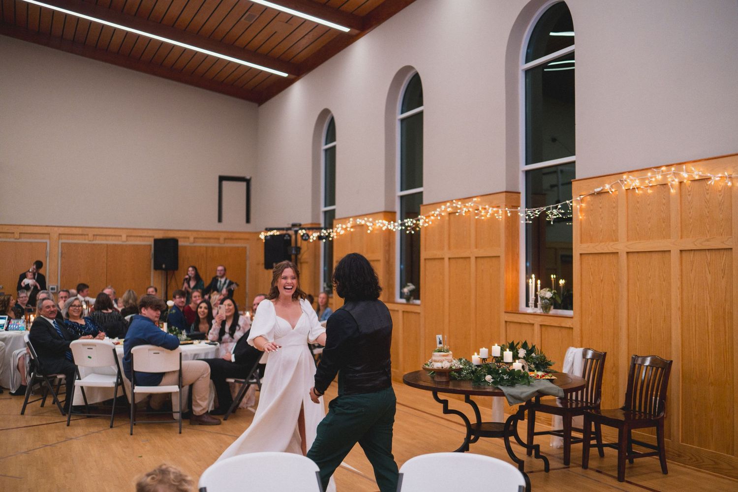 Wedding guests enjoy a celebration in a reception hall with wooden walls and string lights.