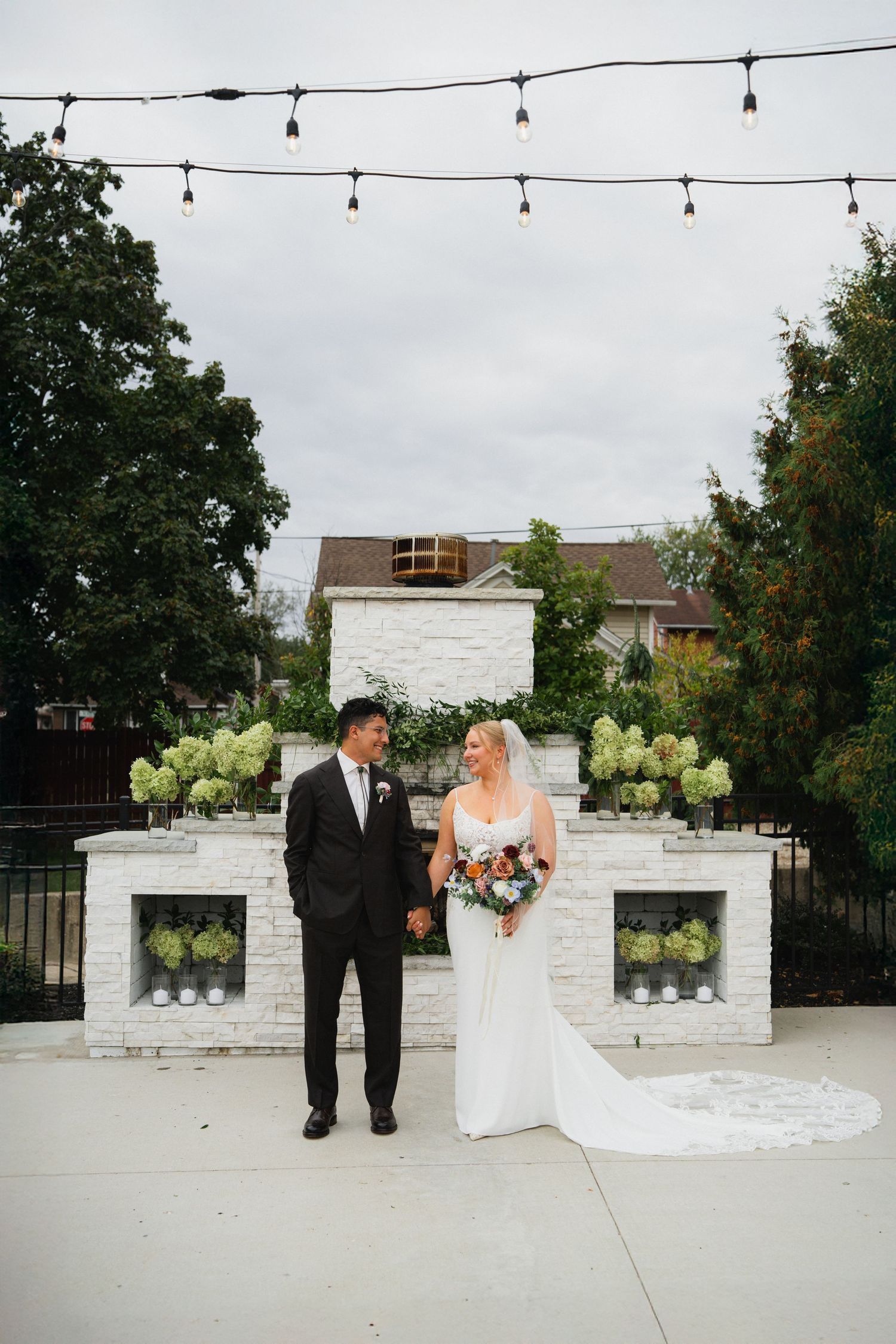 A romantic wedding ceremony takes place at an outdoor venue with white stone pillars and string lights overhead.