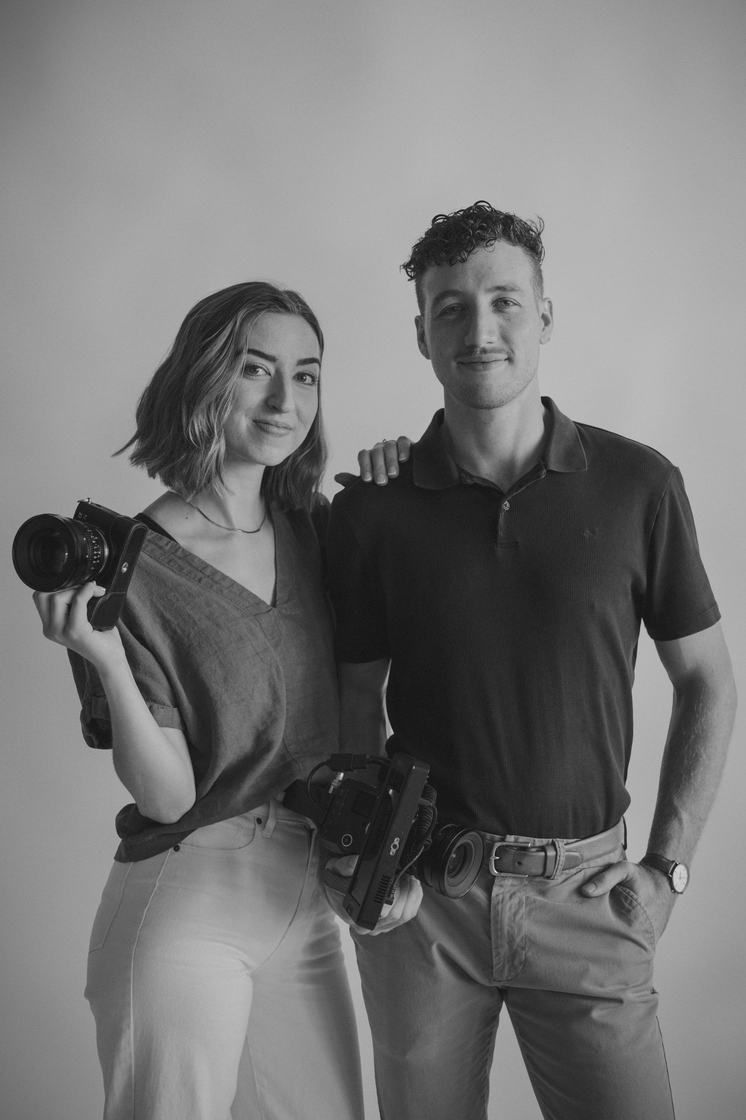 Professional photography and filmmaking duo posing with their cameras in a minimalist studio.