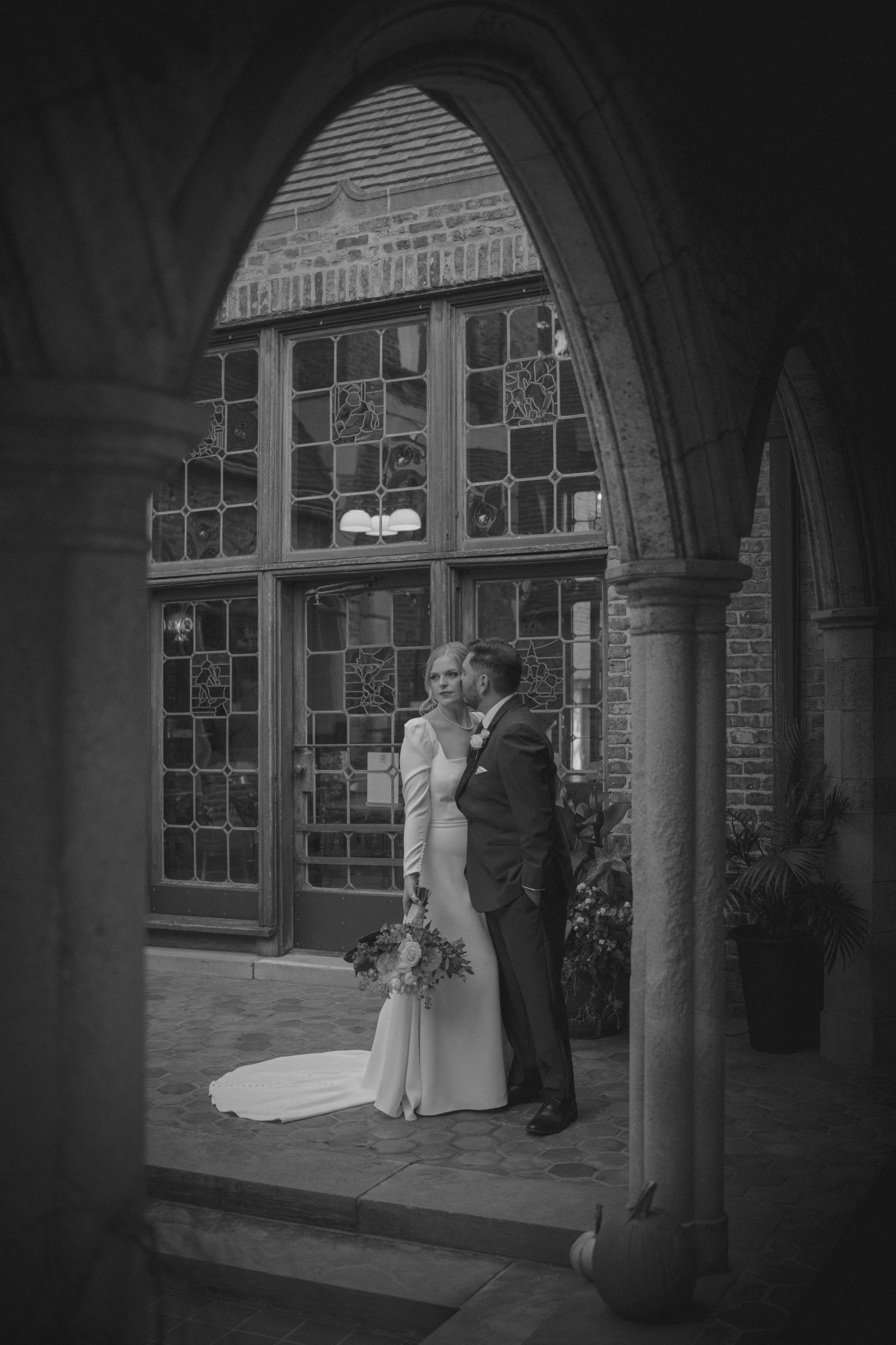 A romantic wedding couple embracing under a gothic stone archway with stained glass windows.