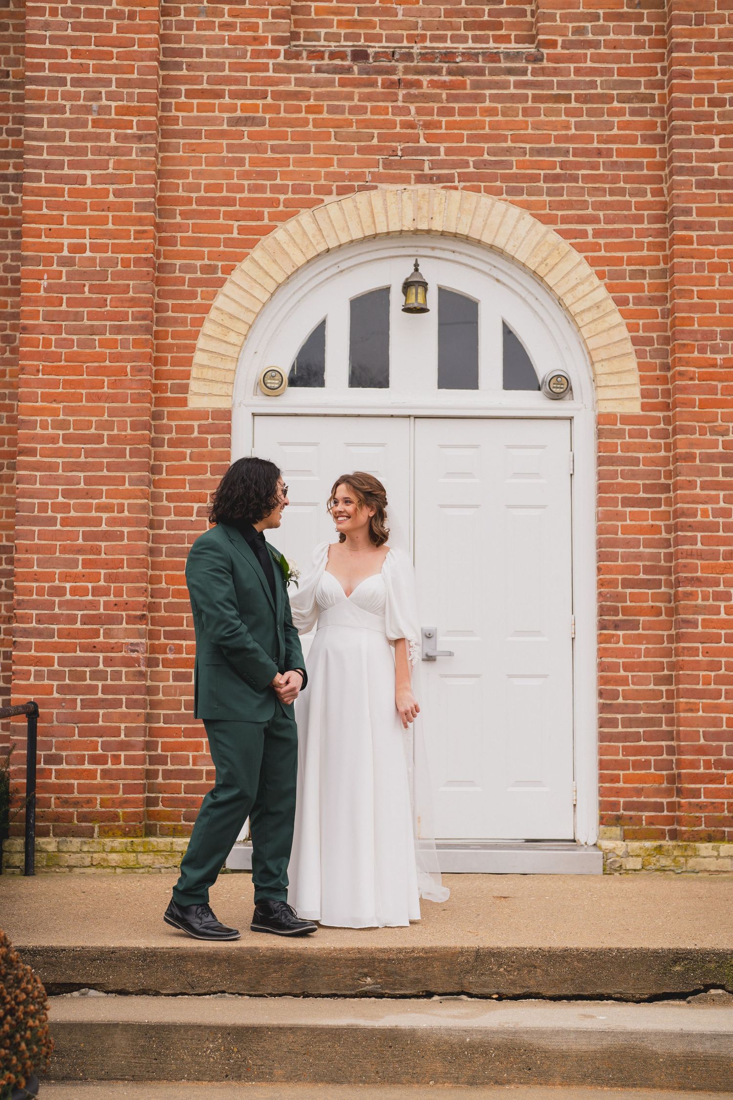 Wedding couple poses playfully in front of a historic red brick church entrance.