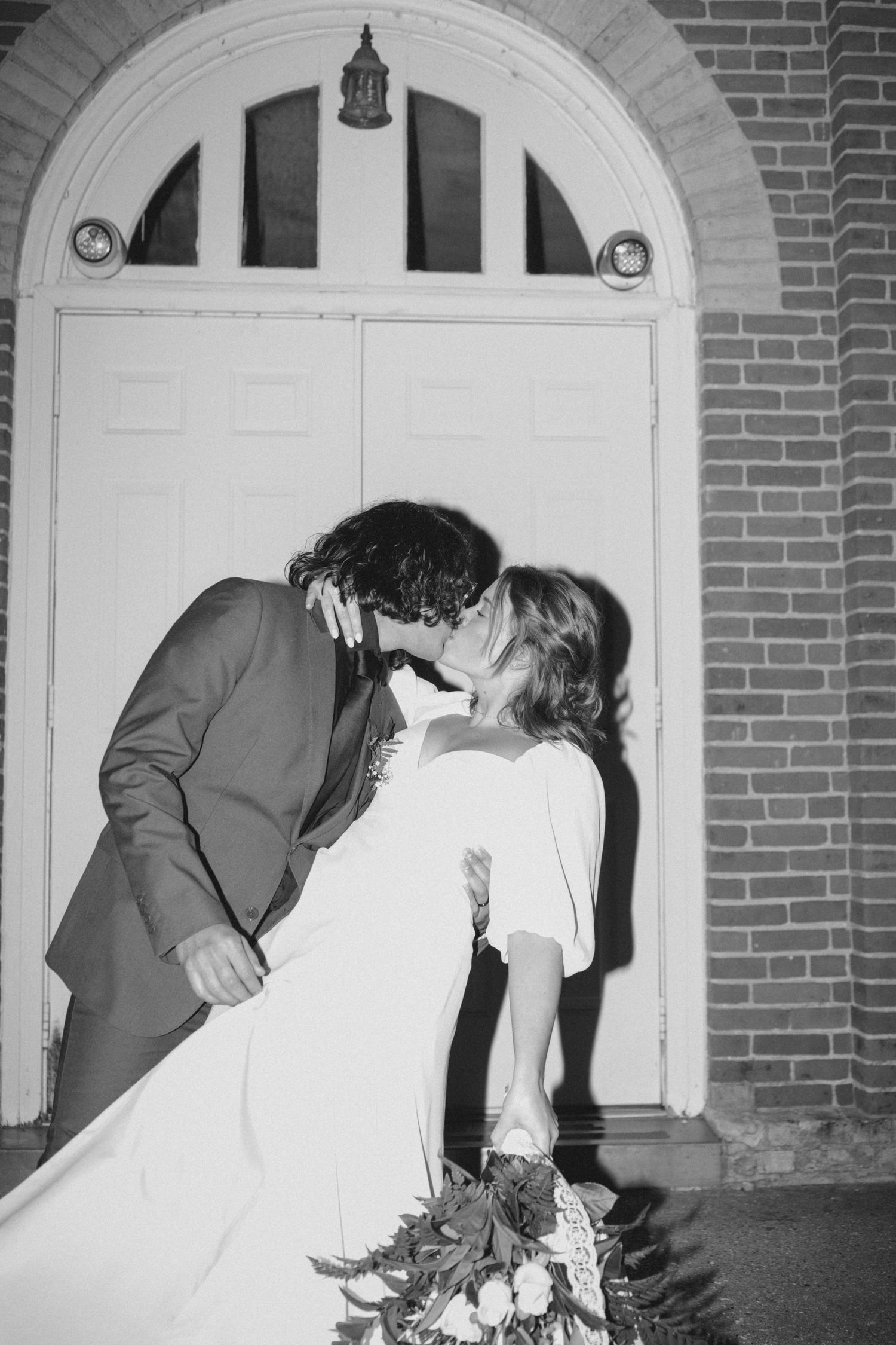 A romantic wedding couple shares a dramatic dip kiss in front of a brick building with white doors.