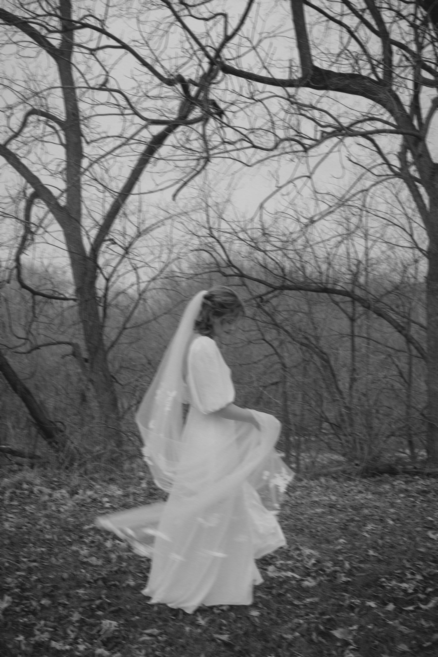 Black and white photo of a bride's dress flowing in the wind among bare winter trees.